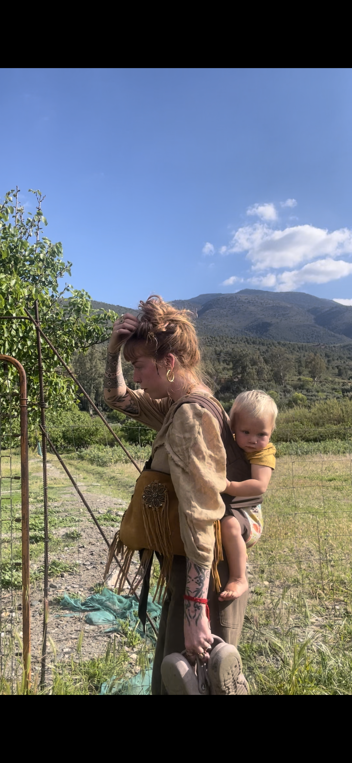 A woman carrying a child on her back outdoors in a rural area with mountains in the background, green fields, a wire fence, and a clear blue sky with clouds.