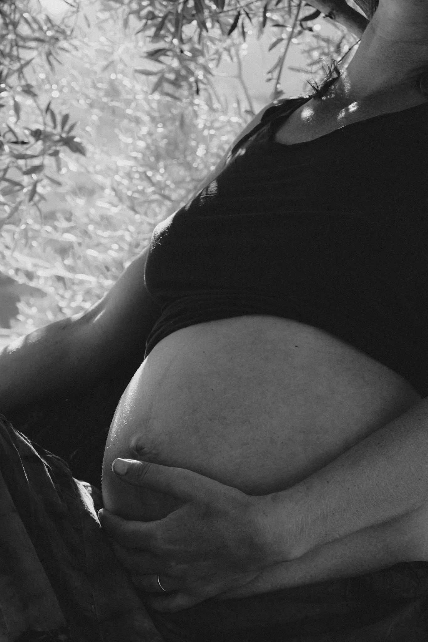 Close-up of a pregnant woman's belly being gently held by her hand, in black and white, with leafy foliage in the background.