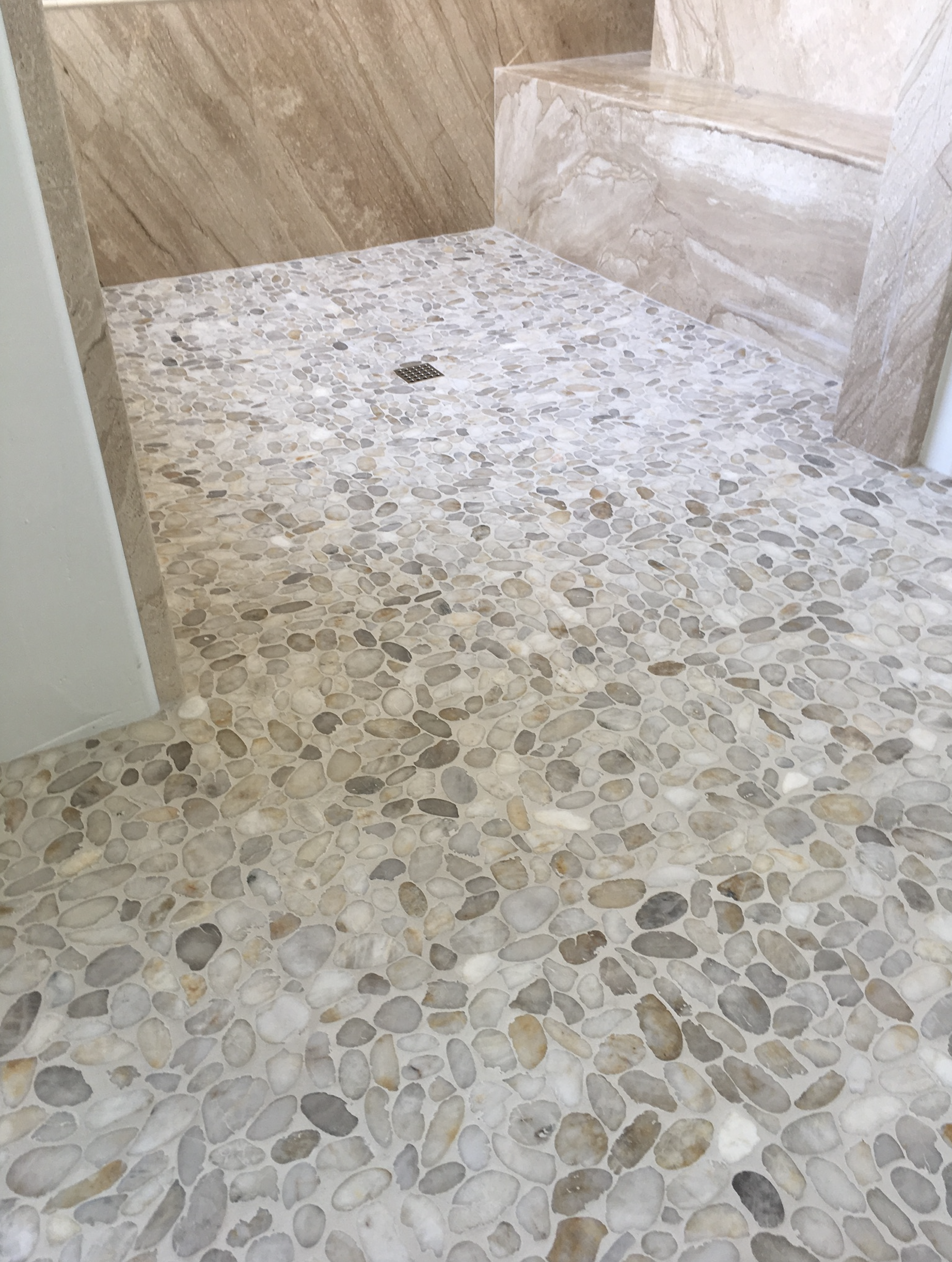 Part of a bathroom shower area with pebble stone floor, a drain in the center, and marble walls and steps.