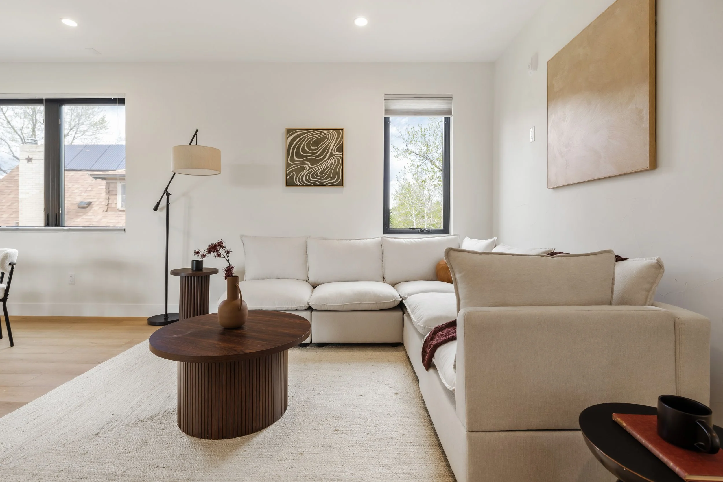 Living room with white sofa, wooden coffee table, abstract art on white walls, window with view of trees, floor lamp, and decorative plant.