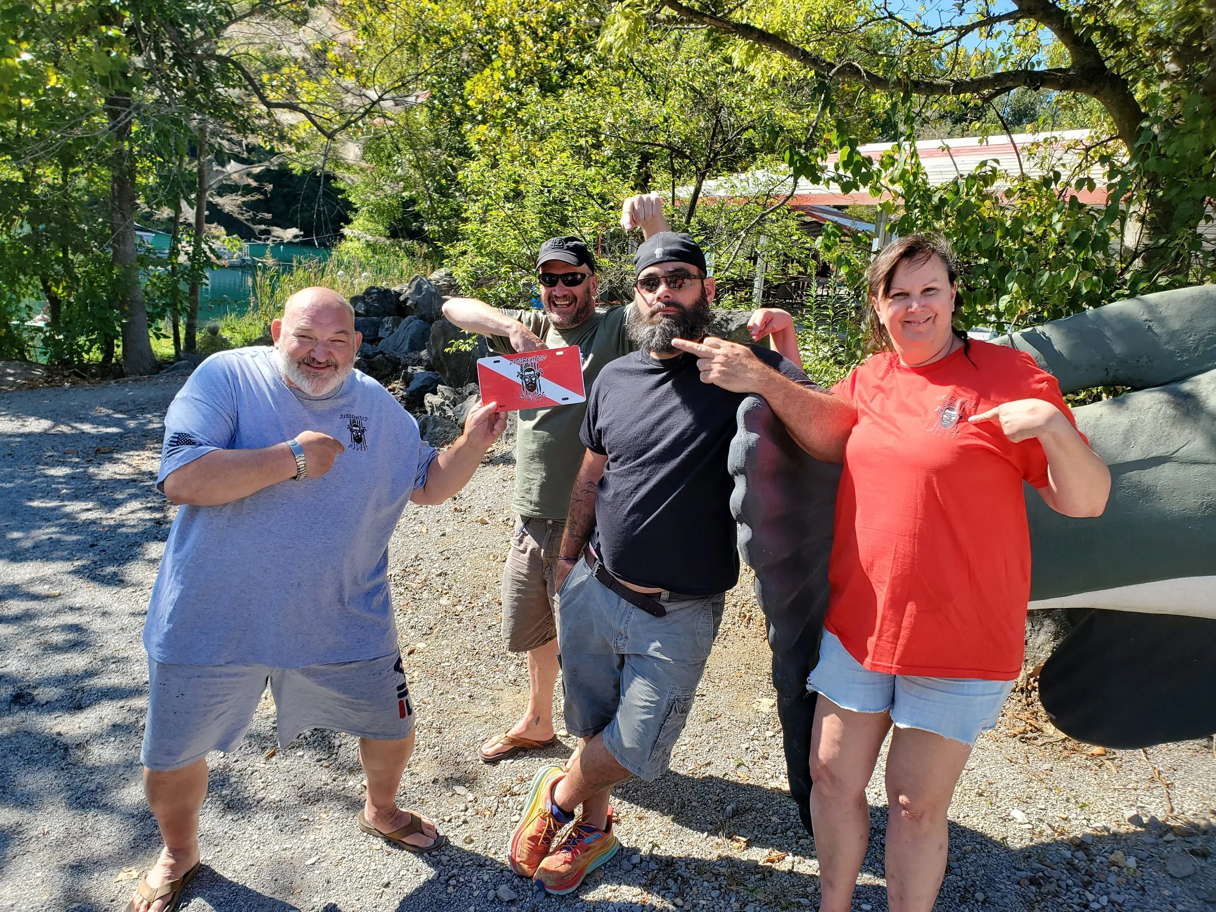 Four people outdoors, three men and one woman, pointing at each other and smiling, with greenery and trees in the background. They appear to be having fun.
