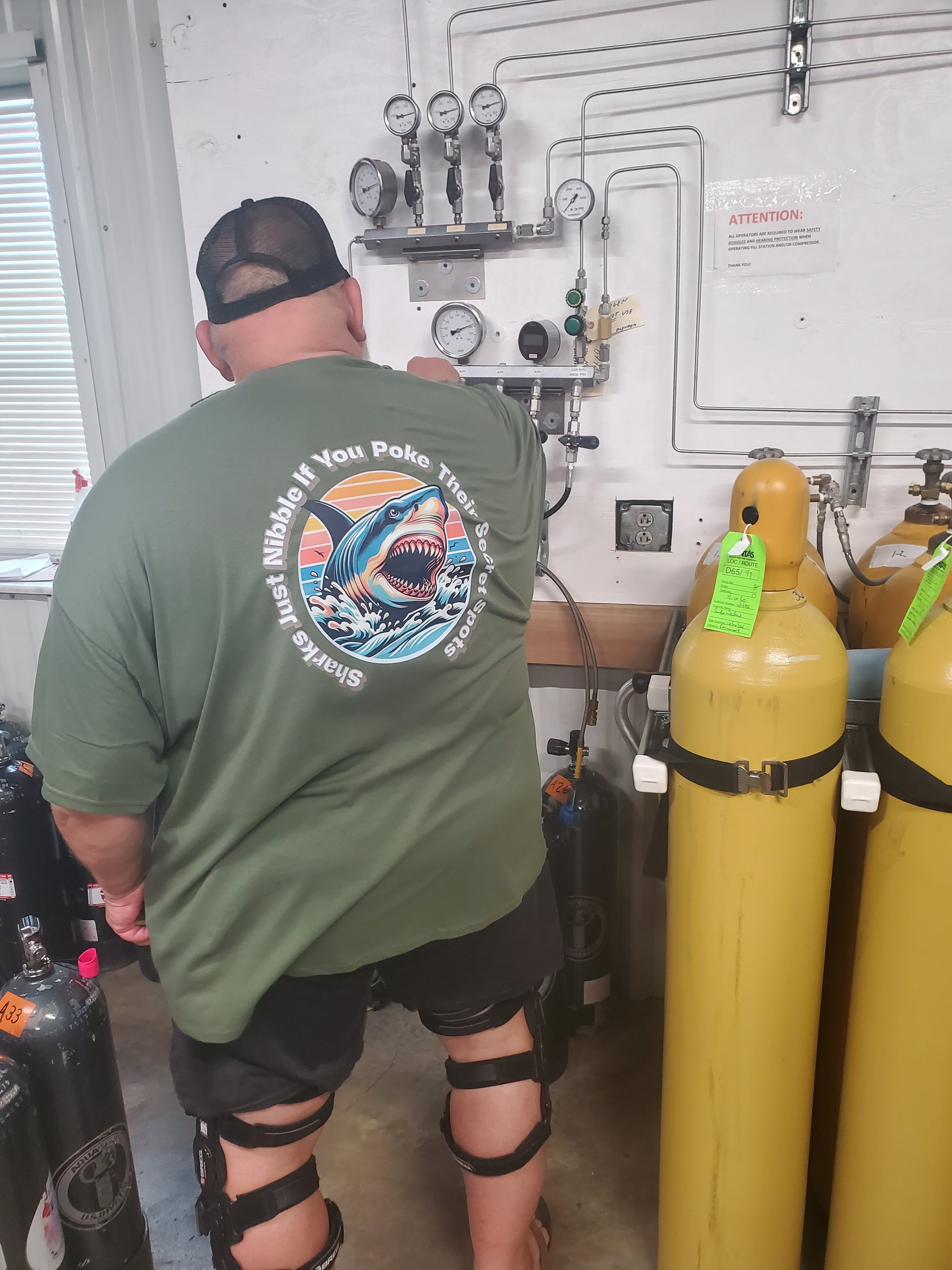 A man wearing a green t-shirt, black shorts with suspenders, and a black cap, working with gas equipment in a technical setting, with gauges and yellow compressed gas cylinders in the background.
