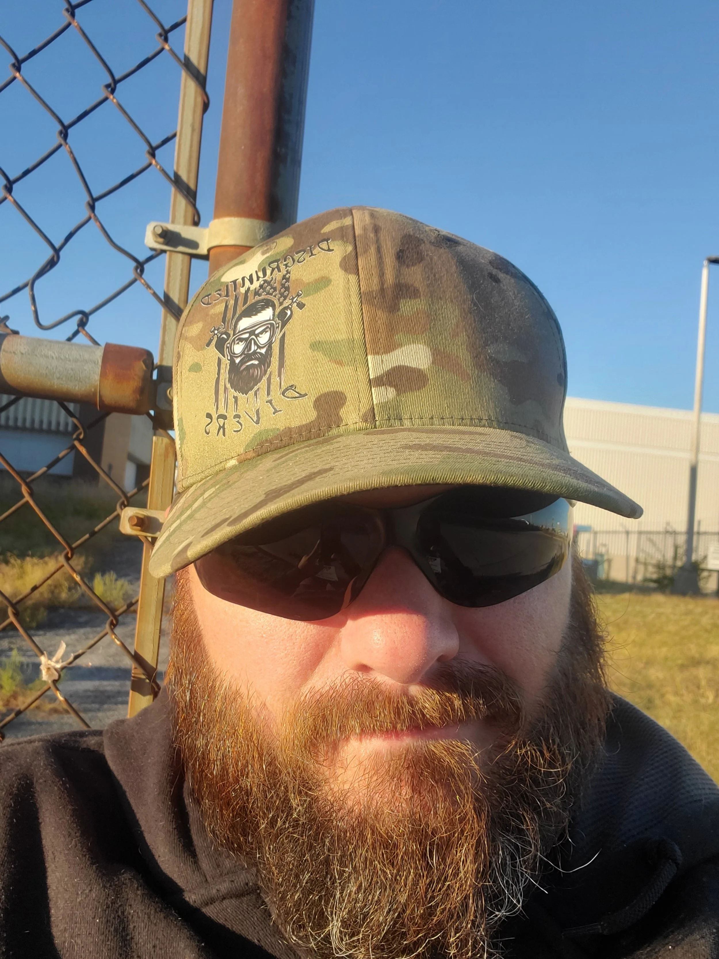 Close-up of a man wearing a camouflage baseball cap with a logo of a bearded man with goggles and headphones, and sunglasses, outdoors beside a chain-link fence with industrial buildings in the background.