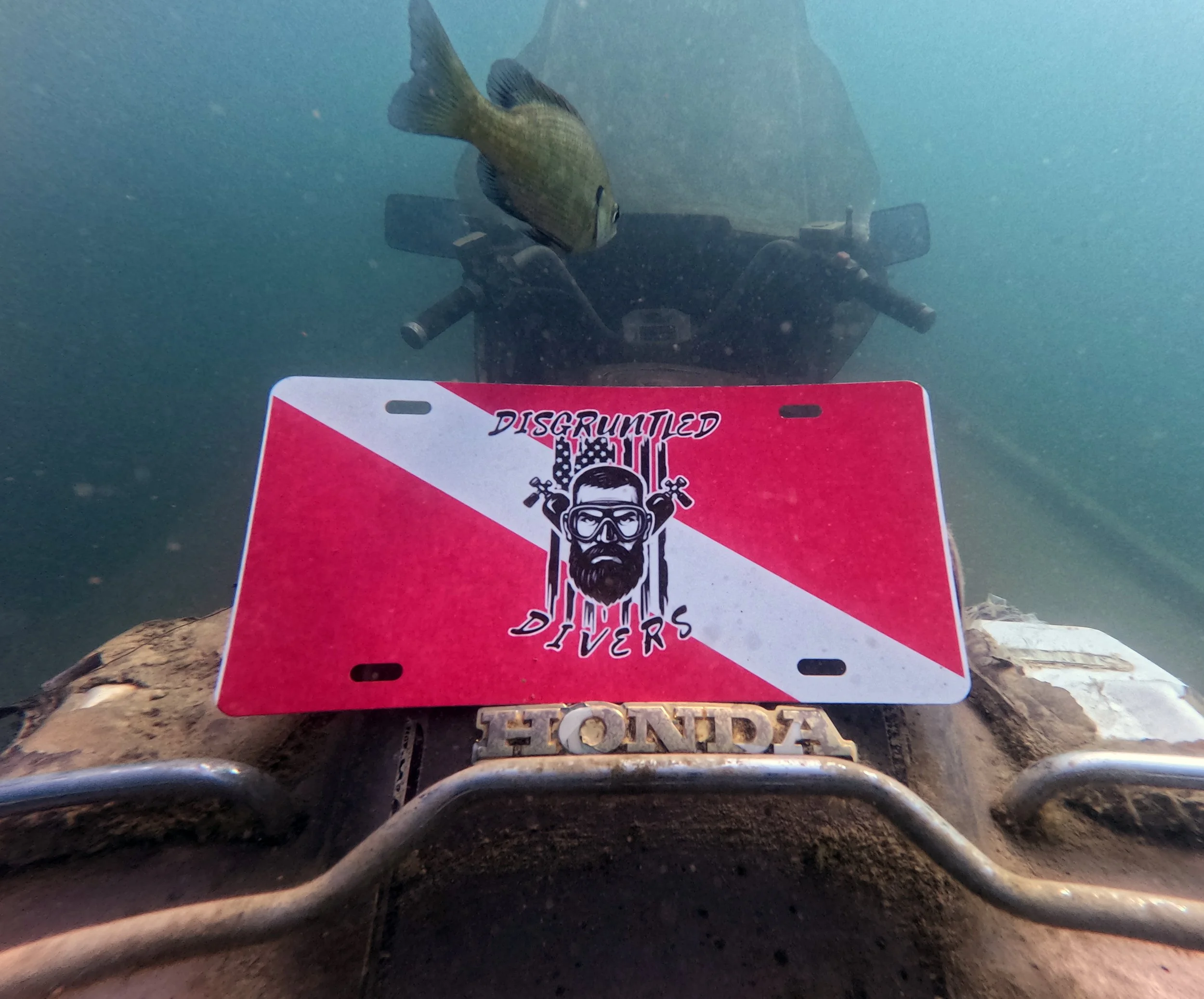 An underwater scene viewed from a submerged Honda all-terrain vehicle (ATV) with a fishing rod and a fish swimming nearby. A red and white sign with the text 'Disgraduated Divers' and a graphic of a diver's face wearing goggles is positioned on the A