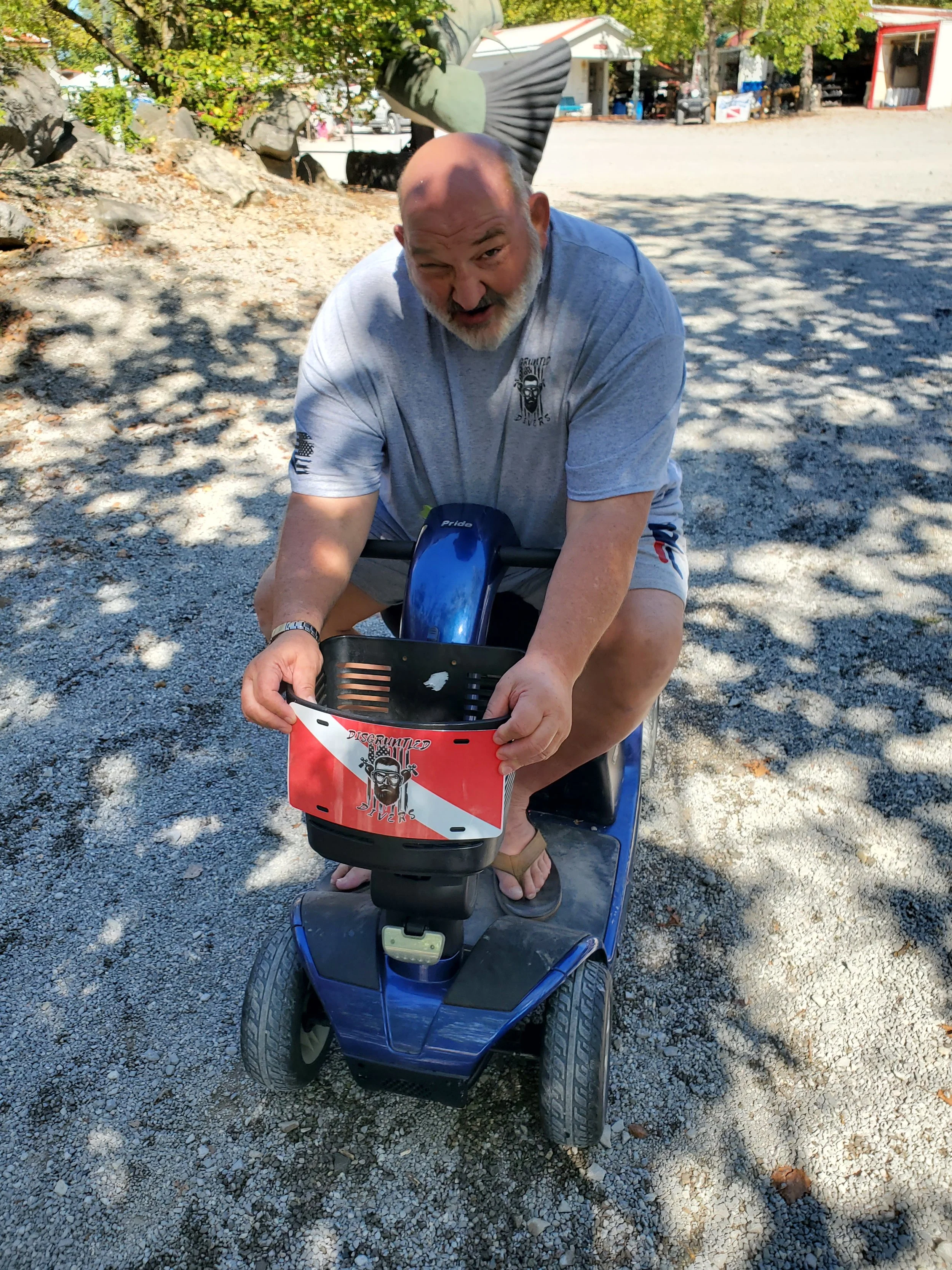 A man sitting on a blue mobility scooter with a red and white logo on the front, outdoors in a sunny area with gravel ground and trees shading him, making a playful face.
