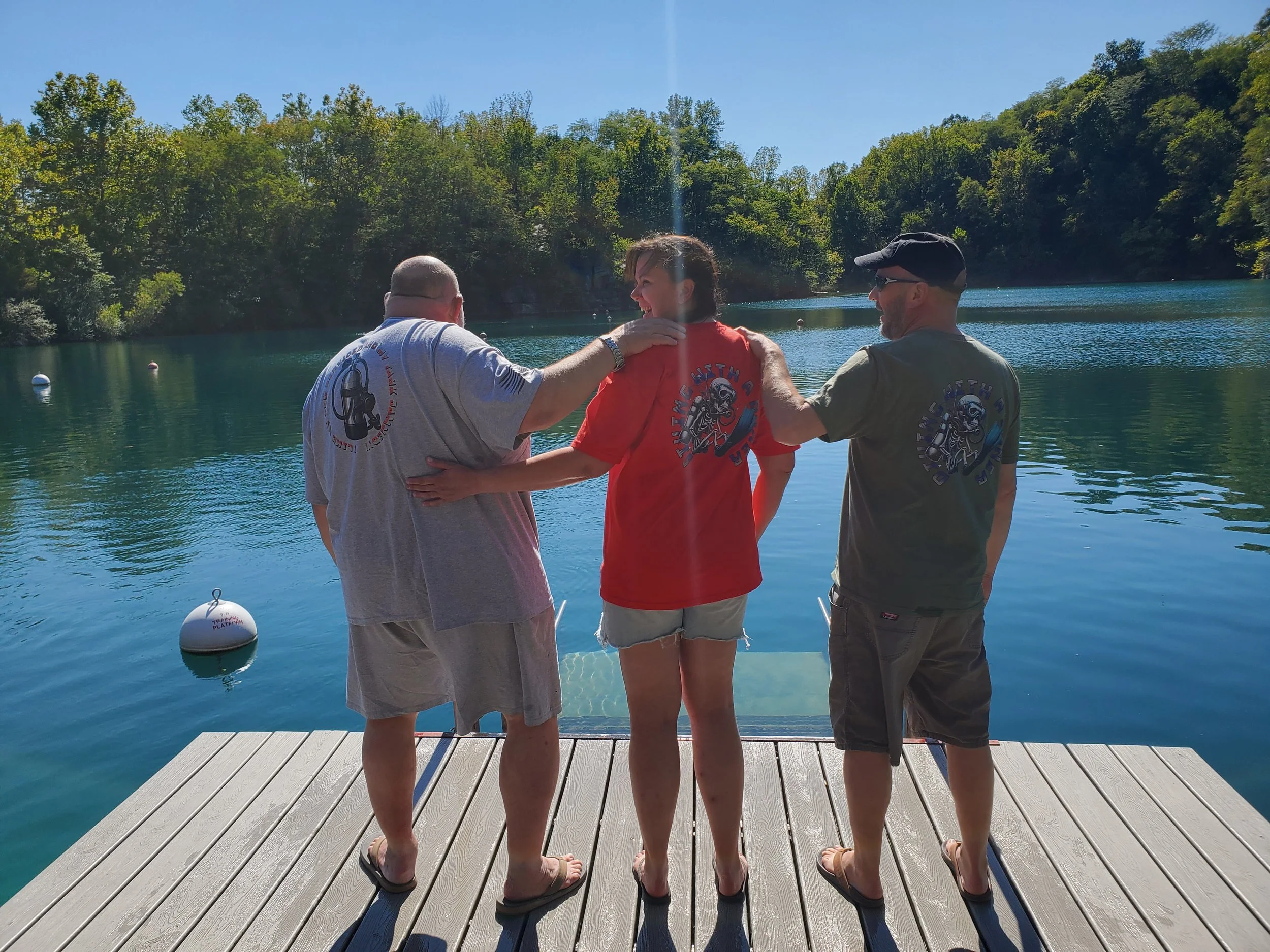 Three friends standing on a dock by a lake, with their backs to the camera. They are wearing t-shirts with a motorcycle logo and are surrounded by trees, under a clear blue sky.