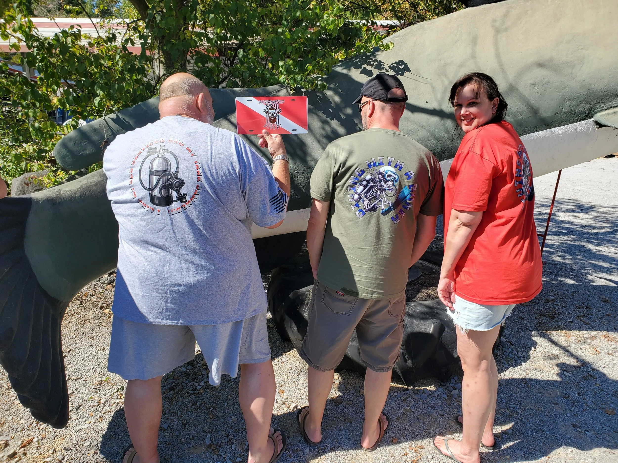 Four people are gathered around a large object, possibly a boat, seen lying sideways outdoors. They are wearing casual clothes with matching event shirts and appear to be examining the object. One person is holding a sign or small board with a graphi