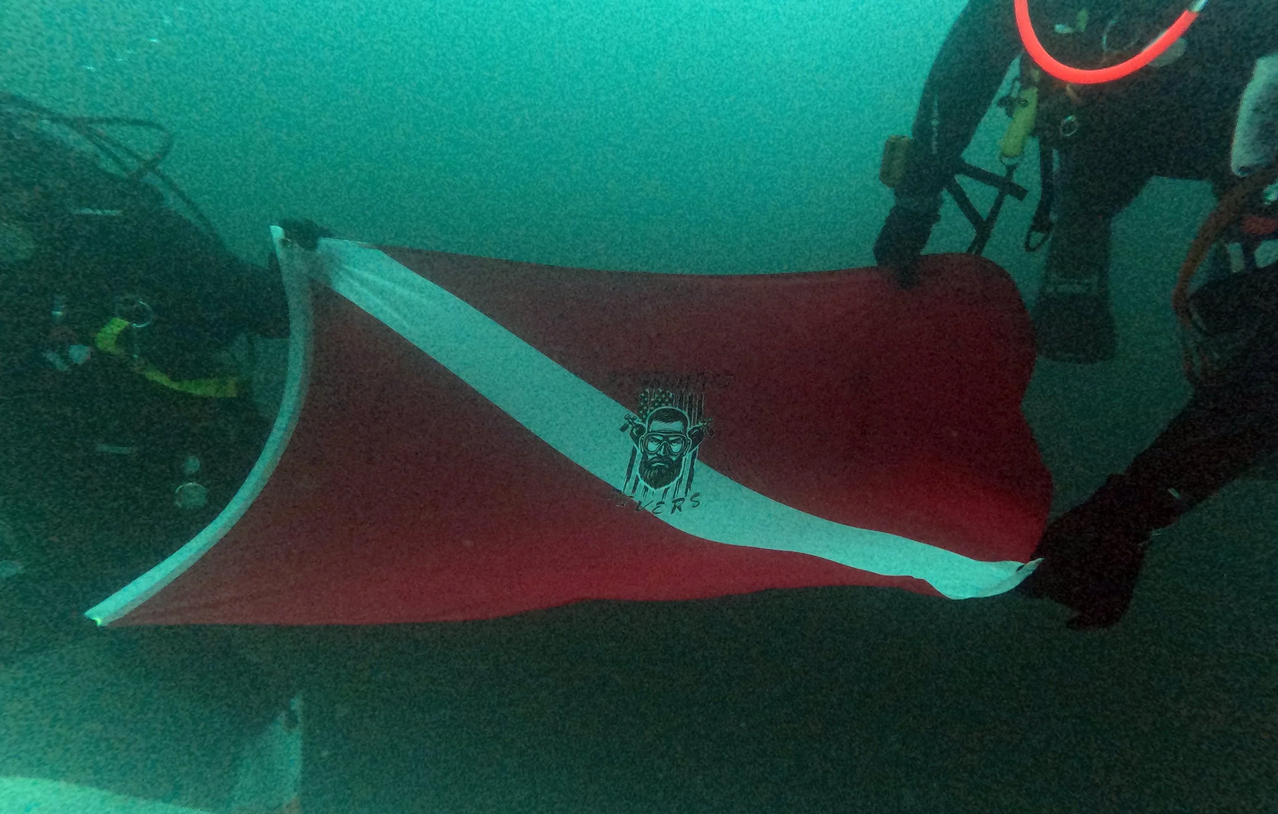 An underwater photograph of a diver holding a red and white flag with a logo of a bearded man with goggles and a crown of tools, surrounded by water.