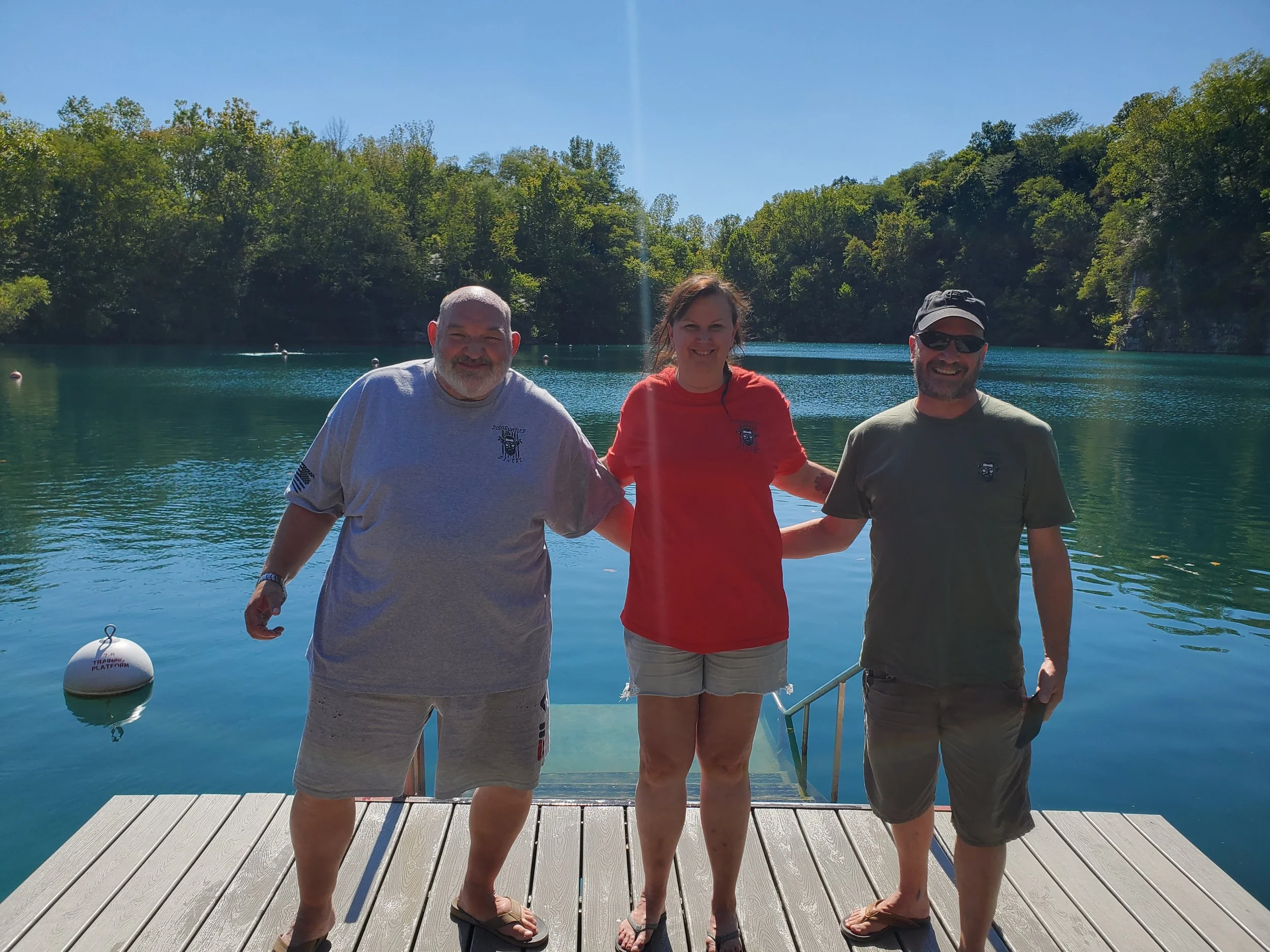 Three people standing on a wooden dock by a lake with green trees in the background on a sunny day.
