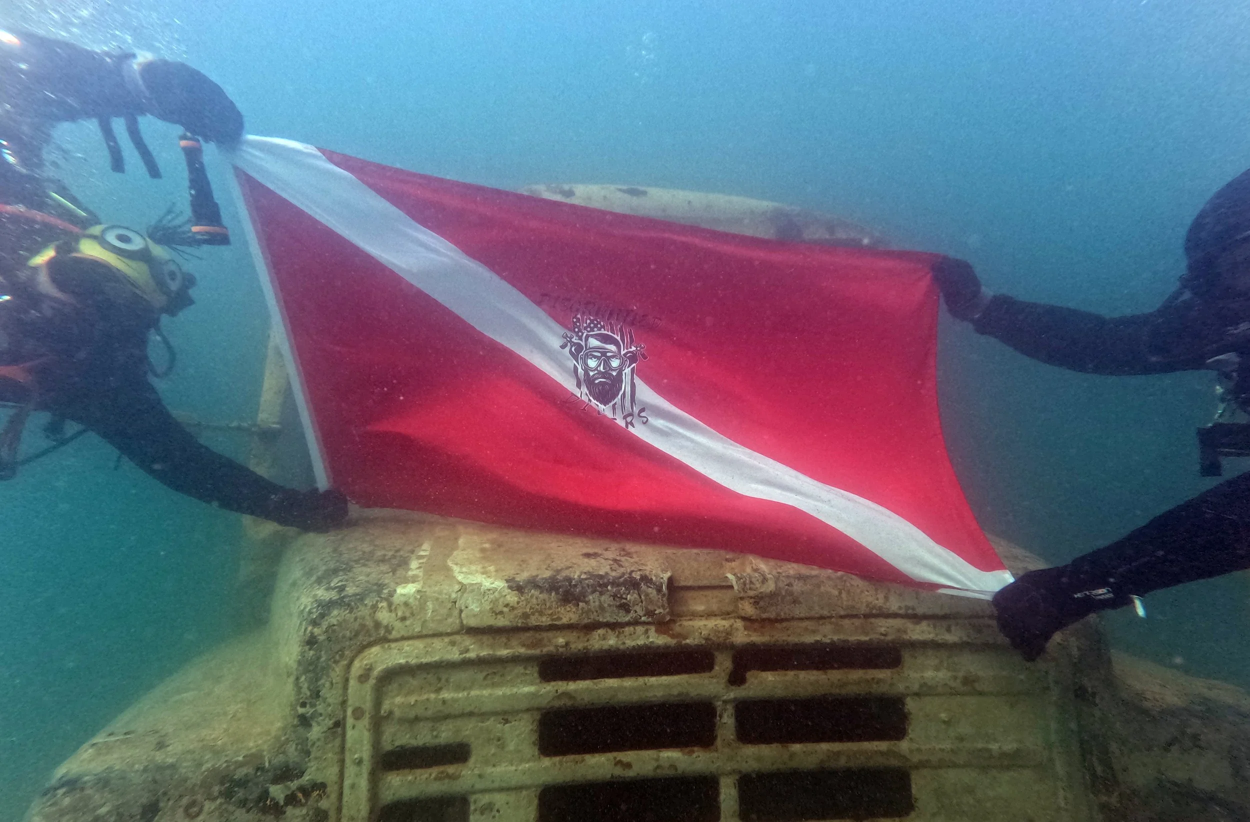Two scuba divers underwater holding a red flag with a white diagonal stripe and a logo of a bearded man with glasses and equipment in the center, atop an old, rusted vehicle or structure.