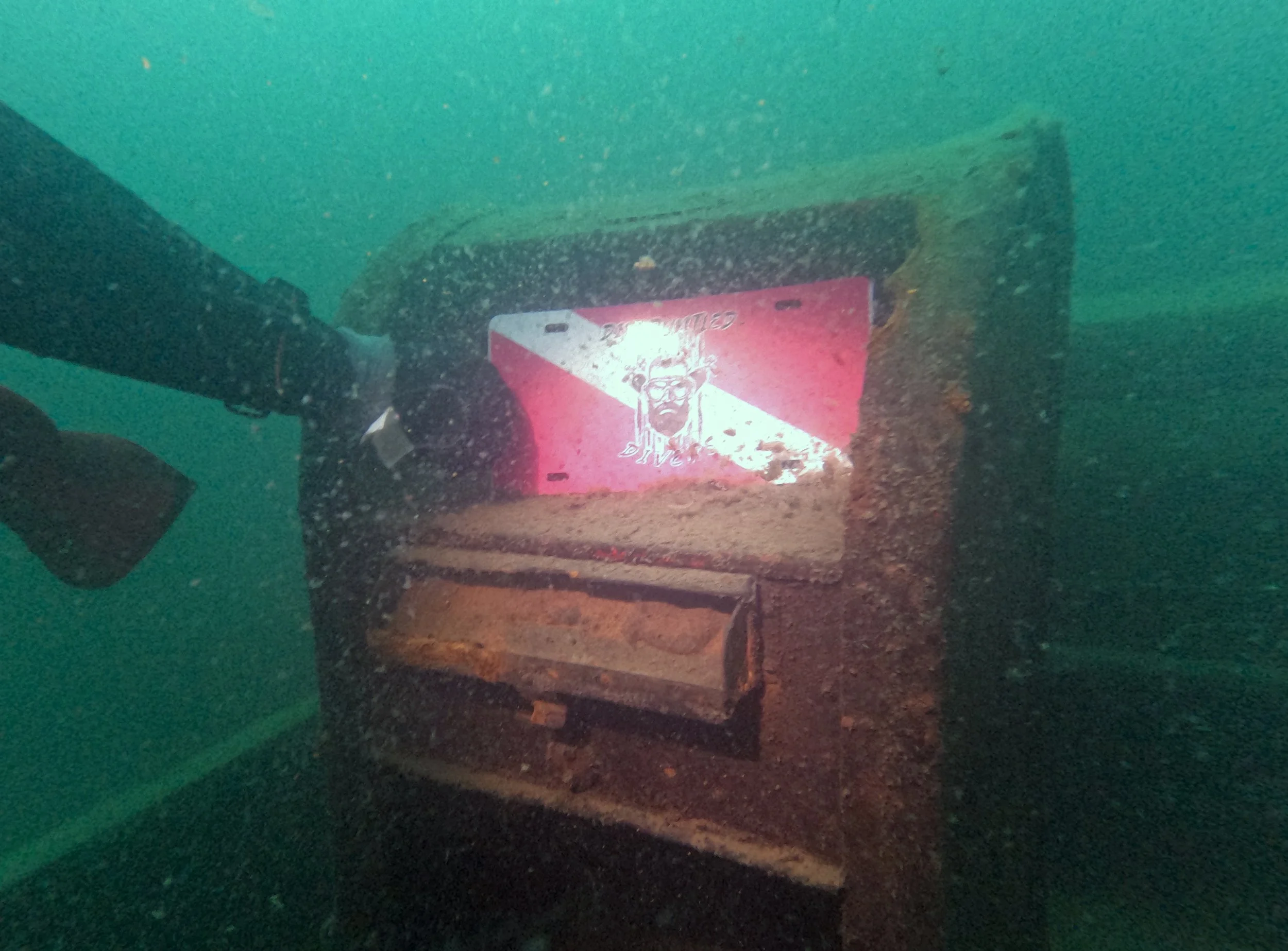 An underwater photograph showing a rusty machine with a partially visible vending machine with a pink and white logo of a bull's head. The machine is covered in sediment and surrounded by murky water.
