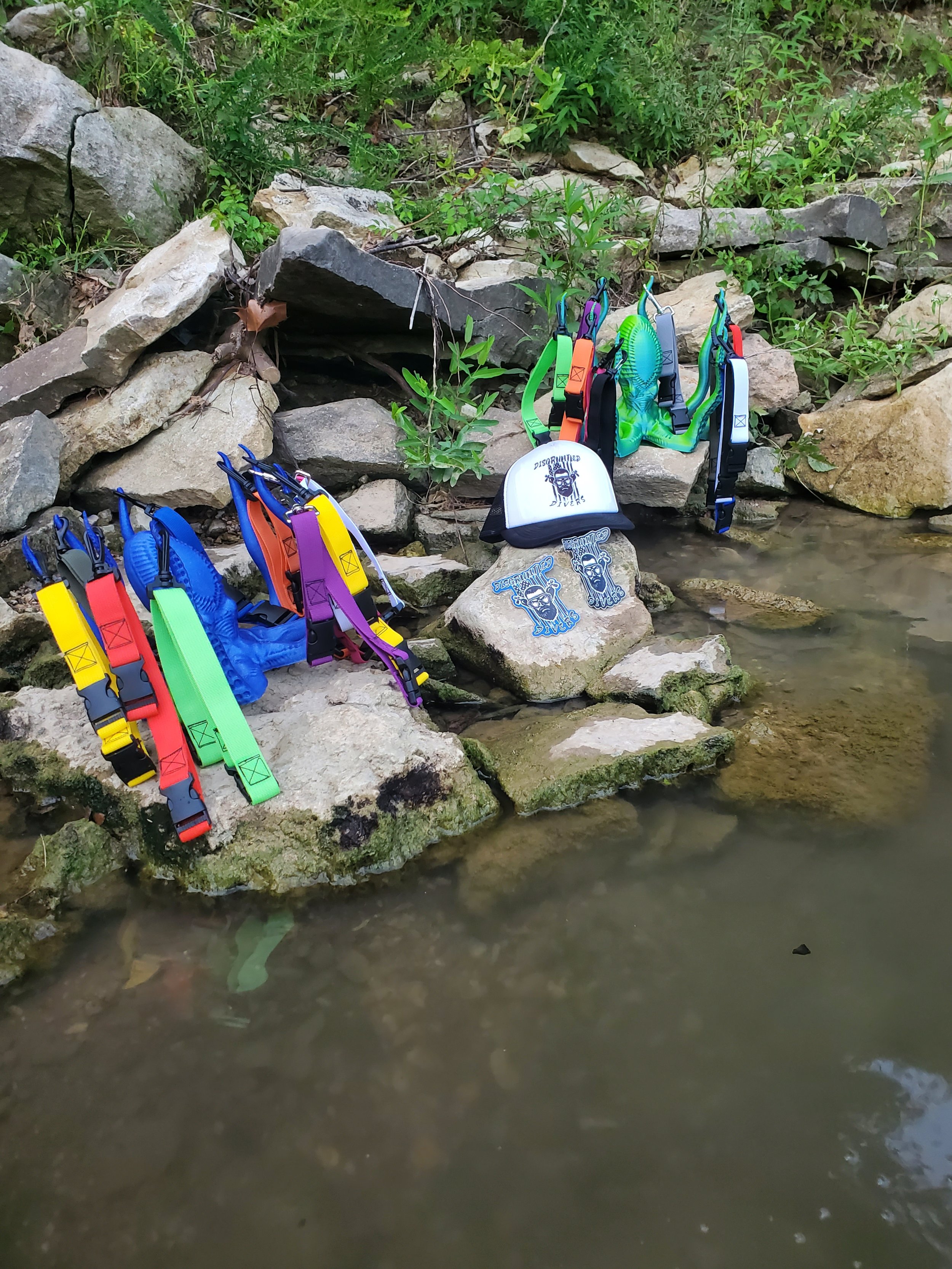 Colorful dog collars and accessories displayed on rocks by a creek surrounded by green plants.