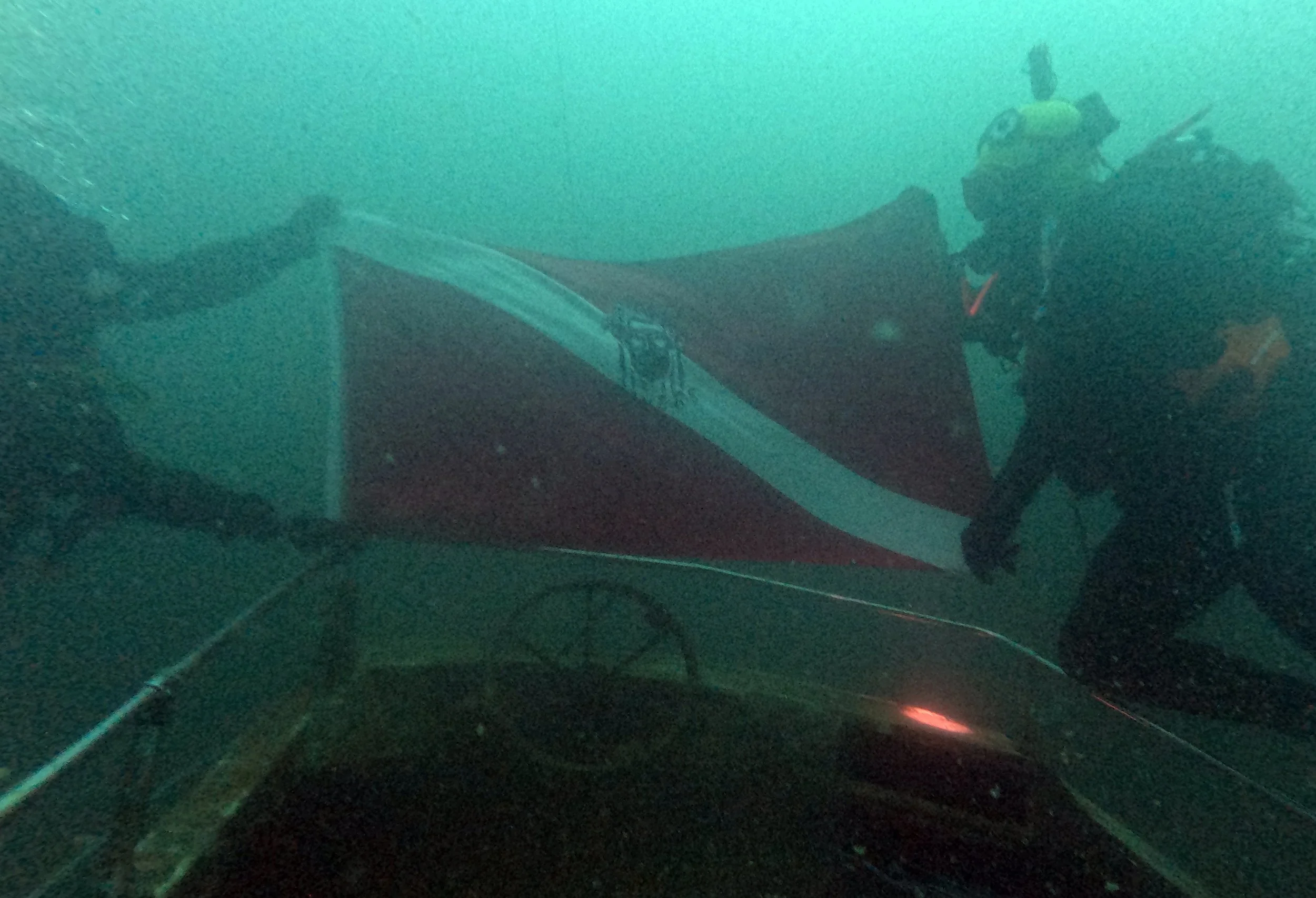 A diver underwater near a boat, with another person's hand and part of the boat visible in the foreground.