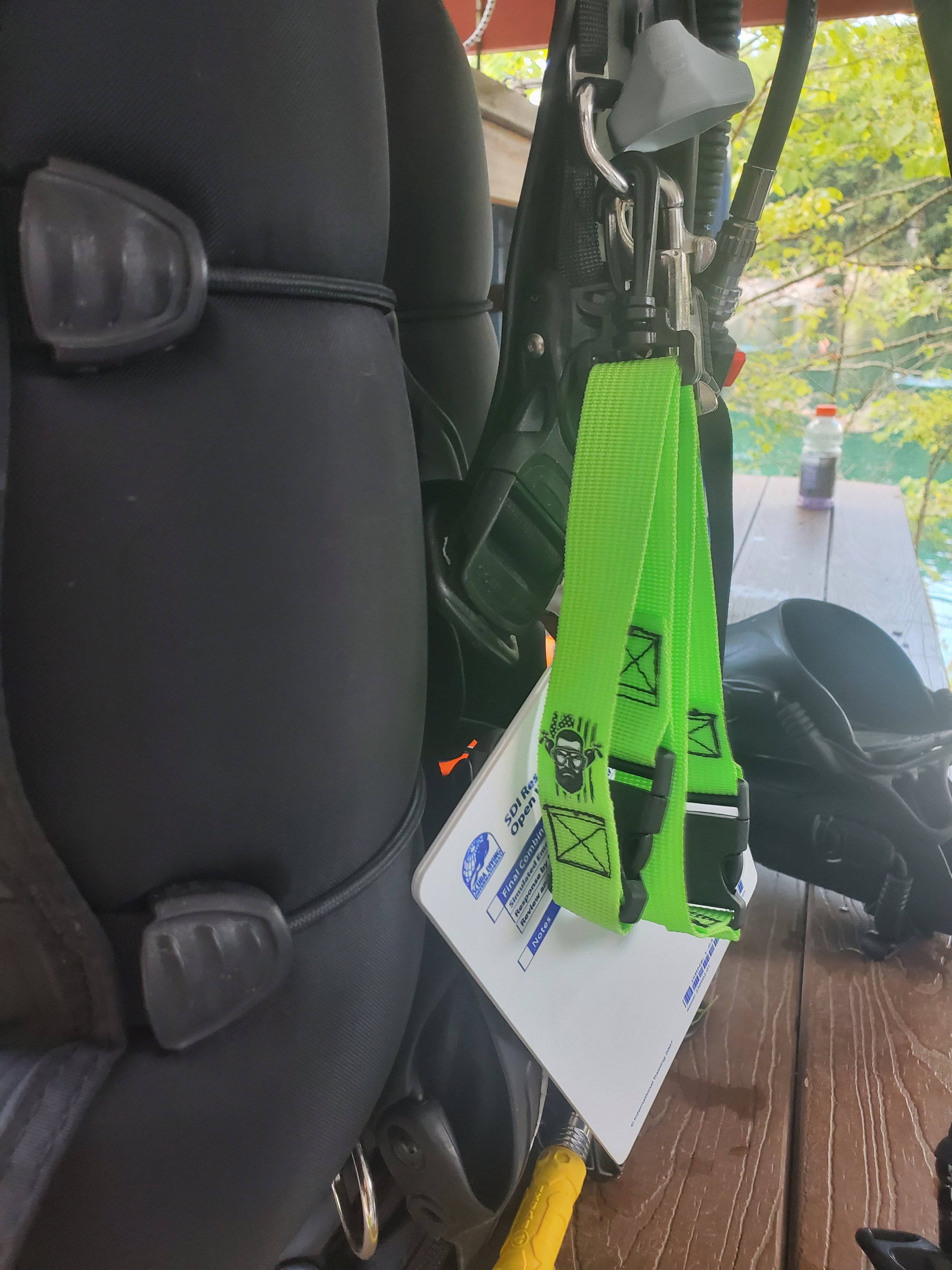 Close-up of a green strap with a black and white logo hanging from the back of a black car seat, with a clip and a white tag attached. There is a black helmet on a wooden surface nearby, and a bottle on the table in the background.