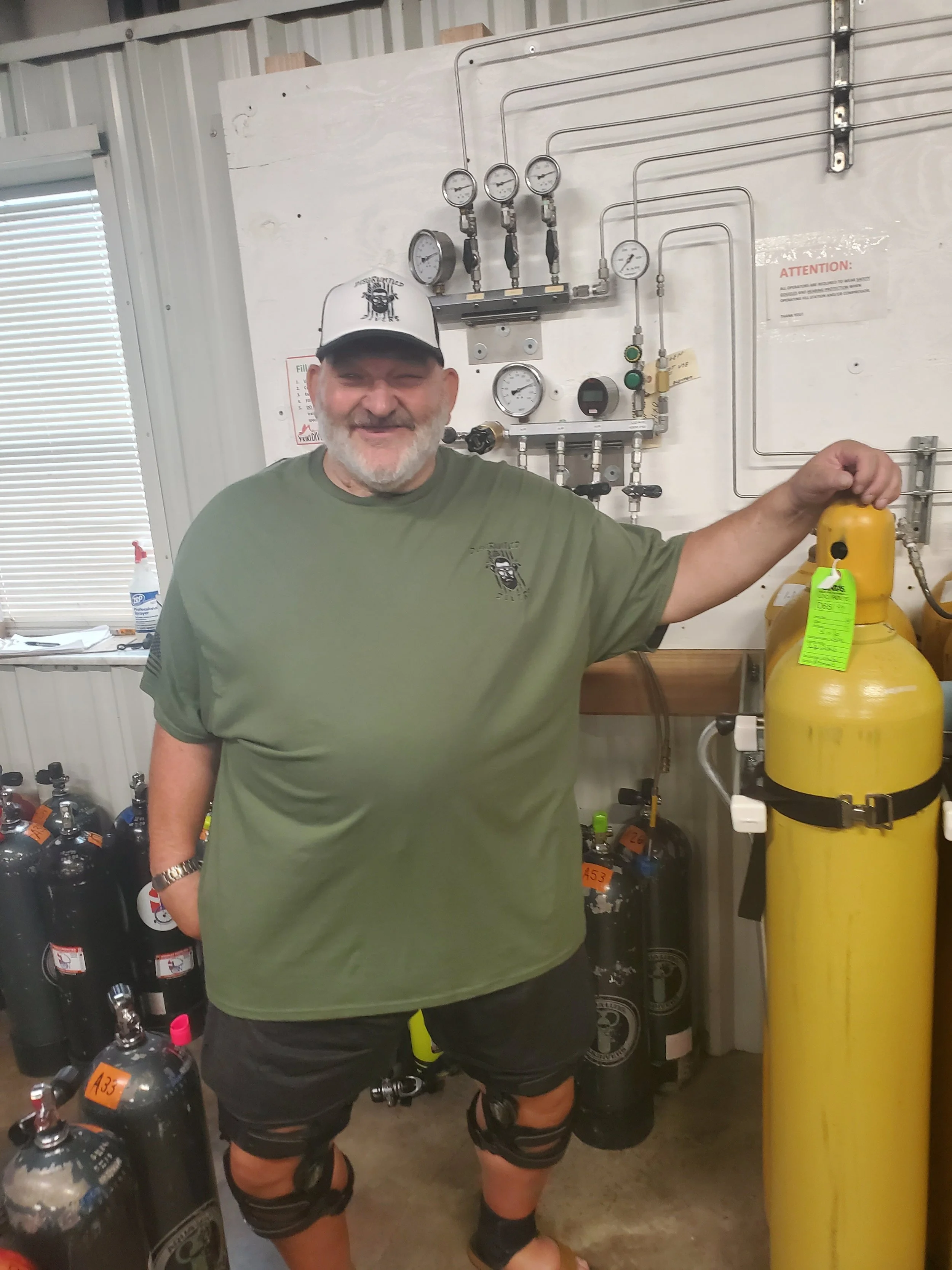 A smiling man with gray hair and beard wearing a green t-shirt, black shorts with knee straps, a white cap, and sandals, standing in a room with diving tanks and gauges, holding a yellow gas cylinder.