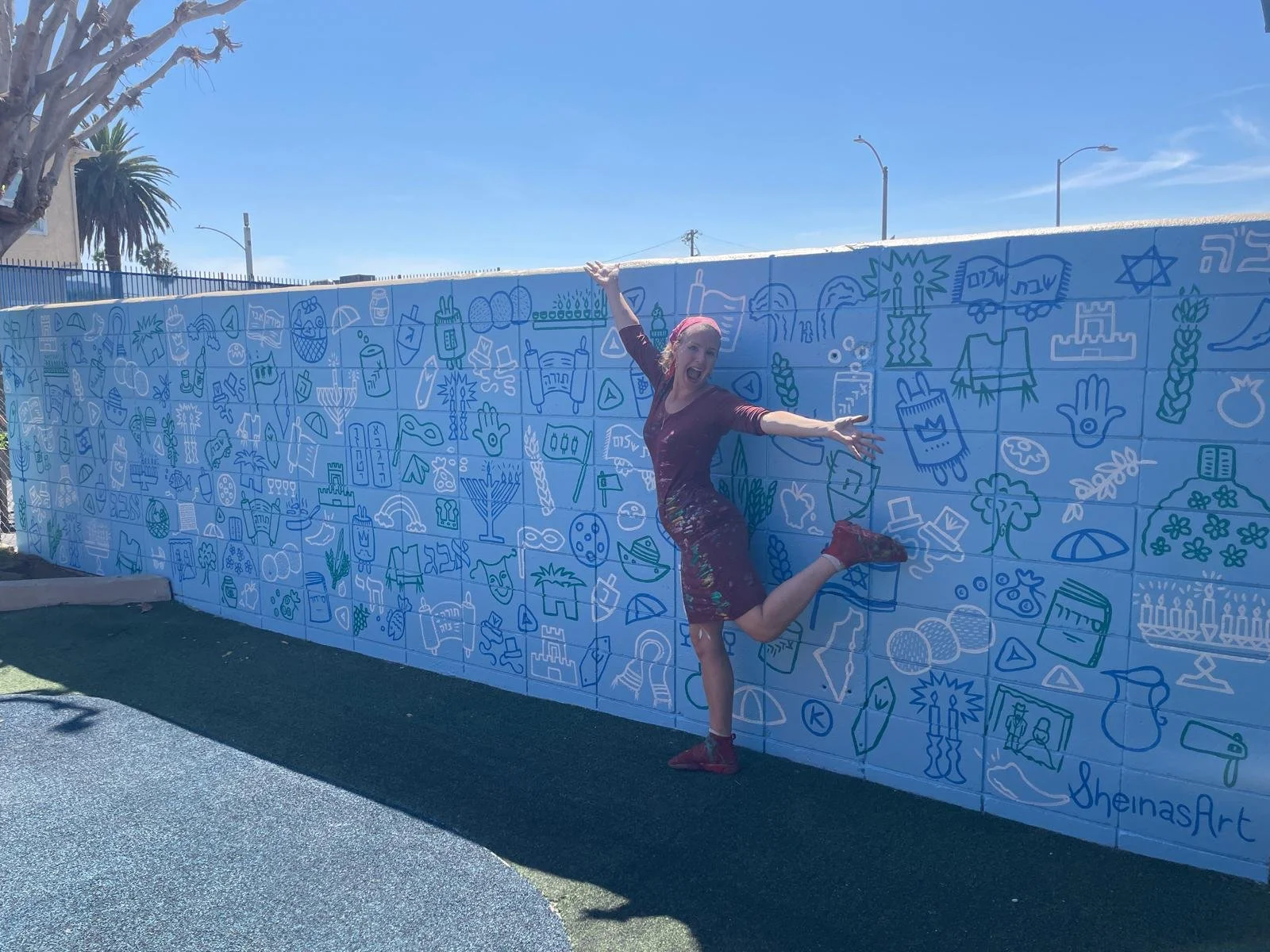 Woman in a maroon dress and red shoes playfully jumps and strikes a pose in front of a colorful mural wall with various objects and symbols, under a blue sky with some palm trees and street lamps in the background.