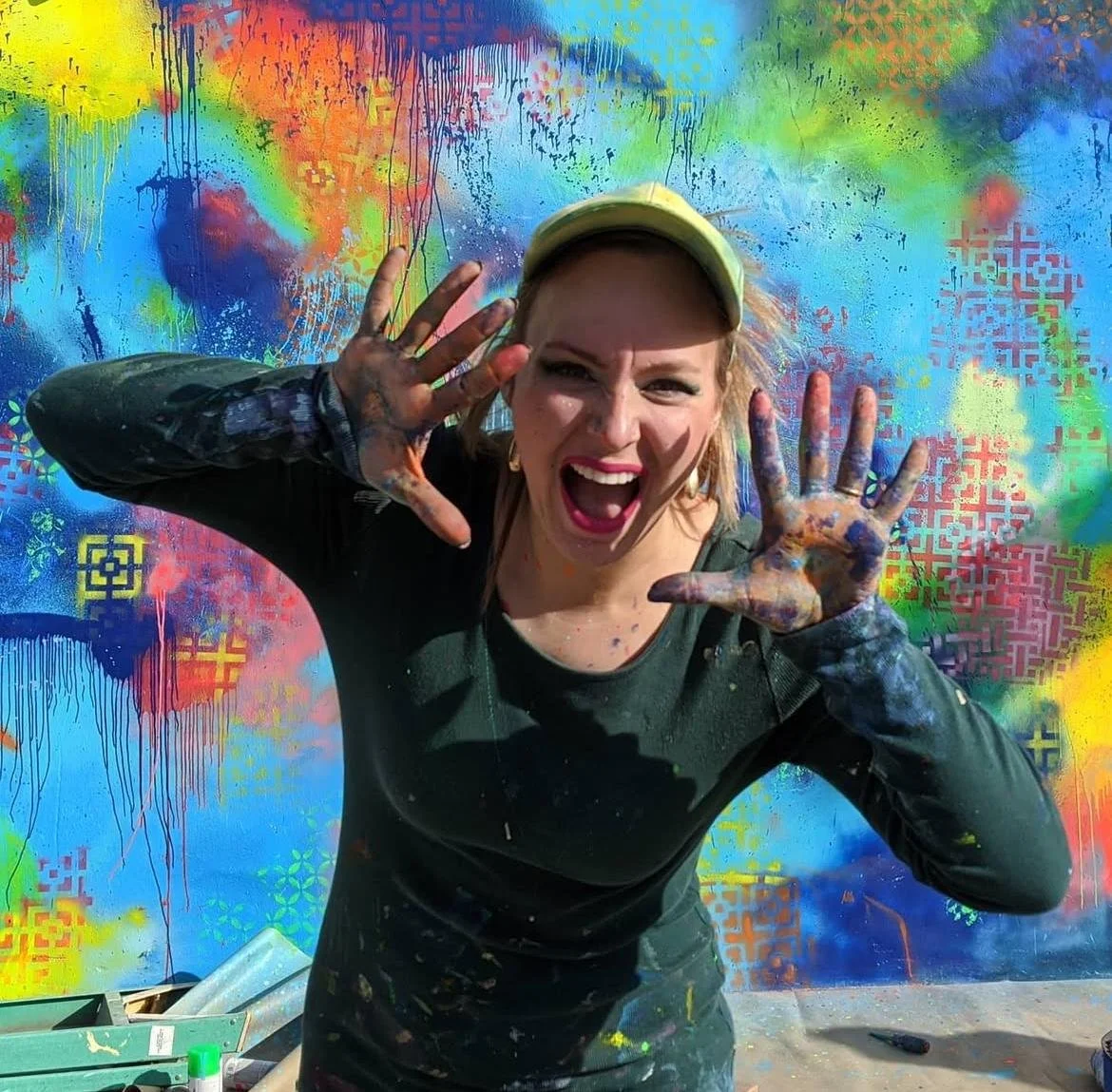 Woman with paint on hands and face, wearing a cap, smiling enthusiastically, standing in front of colorful spray-painted graffiti wall.