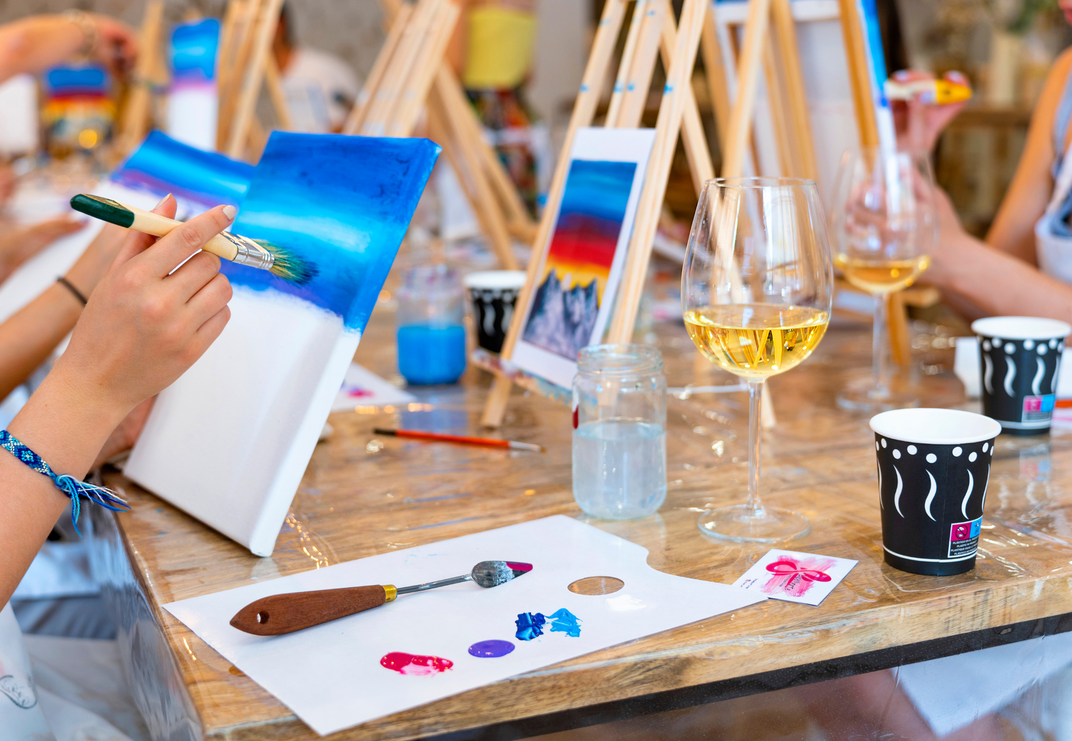 People engaging in a painting activity at a table with canvases, paint, and wine glasses.