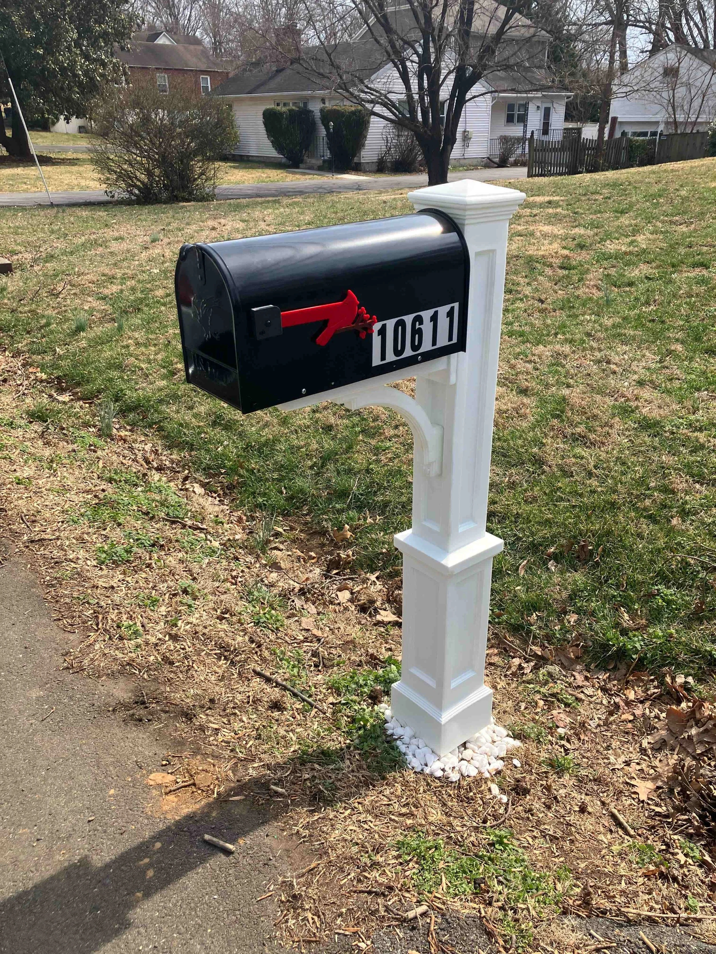 Black mailbox with red flag and white house number 10611 on a white stand, surrounded by small white stones, in a yard with grass and trees in the background.