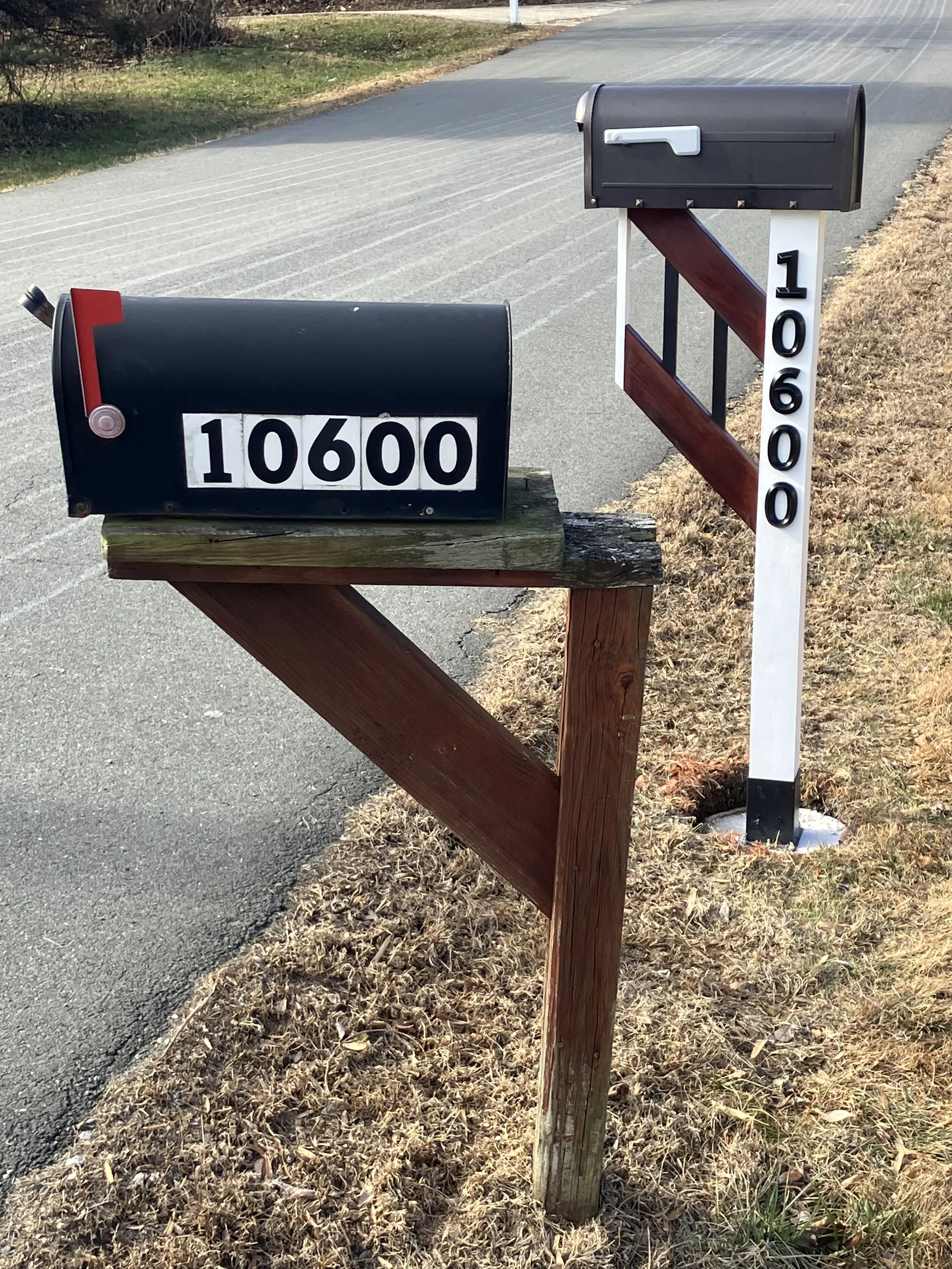 A black mailbox with white numbers displaying 10600 on a wooden post, and a neighboring mailbox with the number 10600 on a white post, next to a sidewalk and brown grass field.