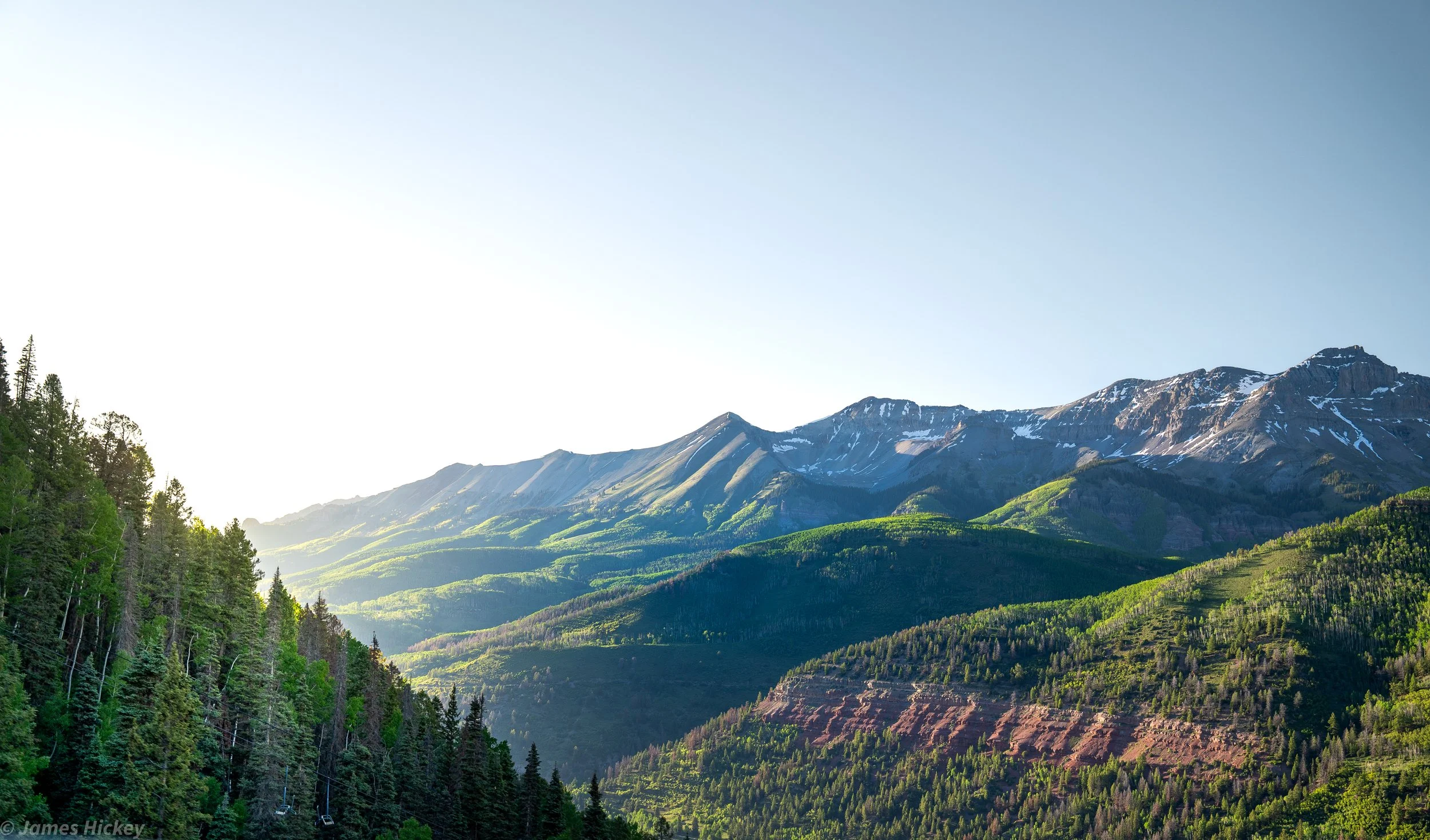 Sunlight illuminating a mountain landscape with snow-capped peaks, green forests, and layered hills.