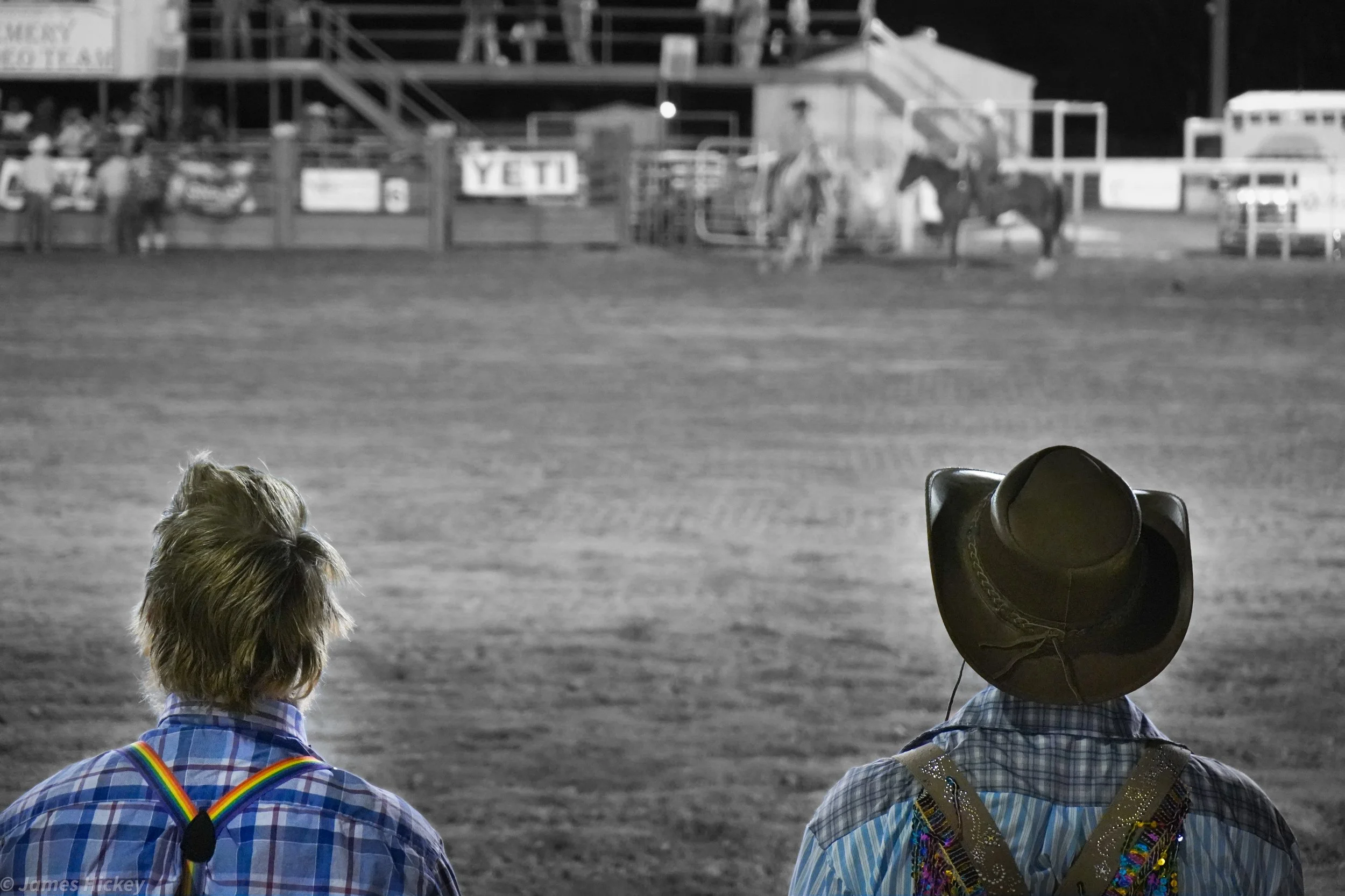 Two people wearing cowboy hats and plaid shirts sitting in front of a rodeo arena at night, with horses in the background.