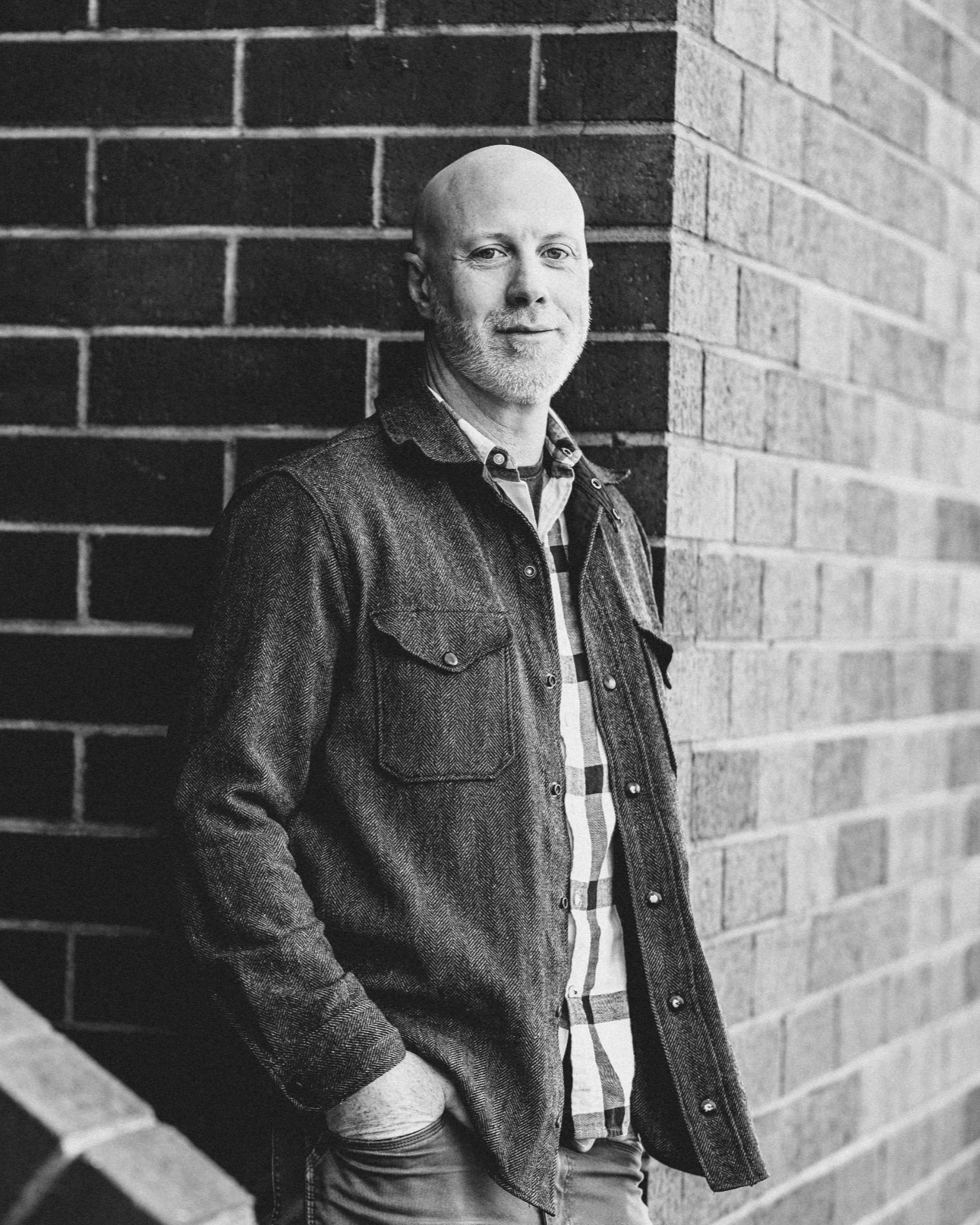 Black and white portrait Evan Hendrix a marriage coach in Leiden, Netherlands. He appears as a bald man with a beard leaning against a brick wall, wearing a denim jacket over a plaid shirt.