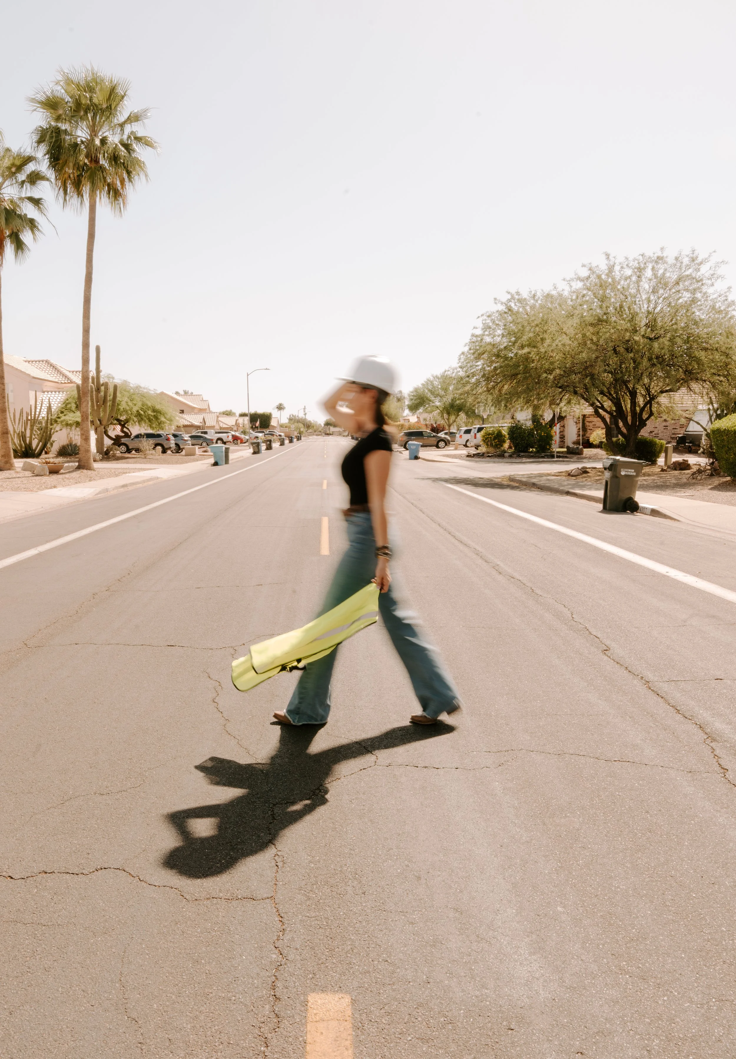 A woman walking across a suburban street wearing a white helmet, black top, and jeans, carrying a yellow bag, with palm trees and houses in the background on a sunny day.