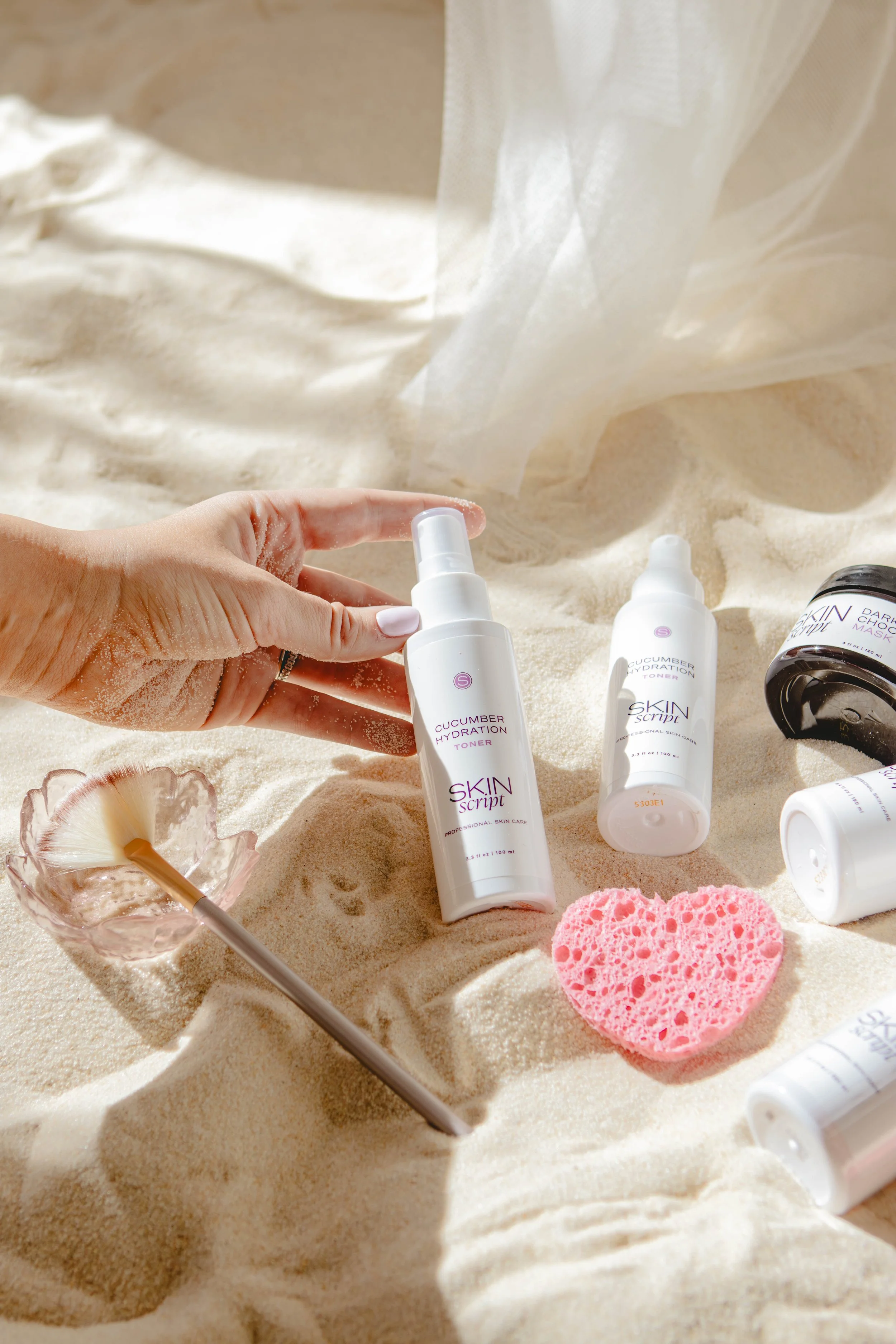 Skincare products on sand, including cucumber hydration toner, pink sponge, and a flower-shaped container with a honey dipper.