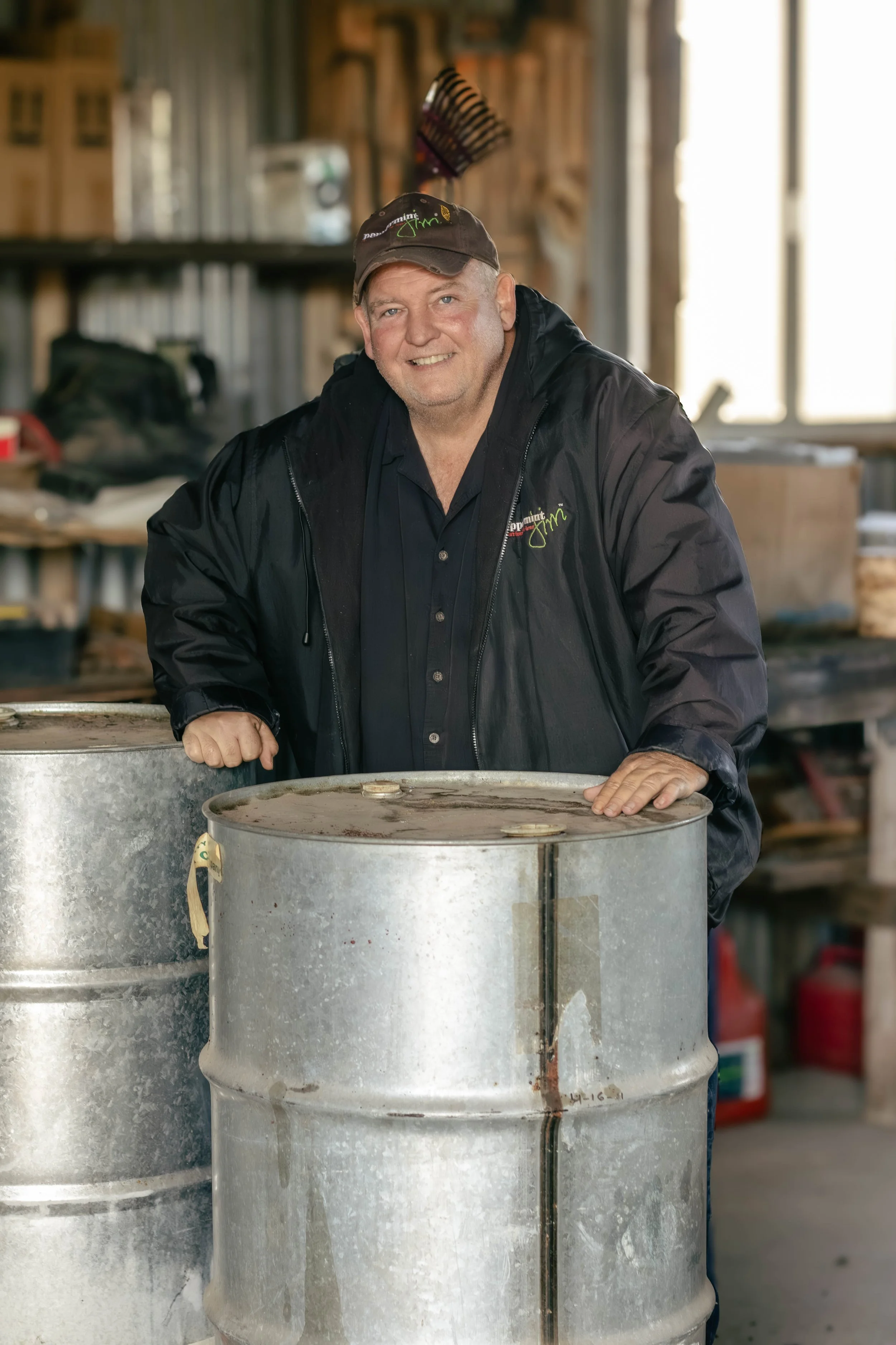 A man in a black jacket and cap smiling, standing behind two large metal drums in a workshop or garage with wooden walls and equipment in the background.