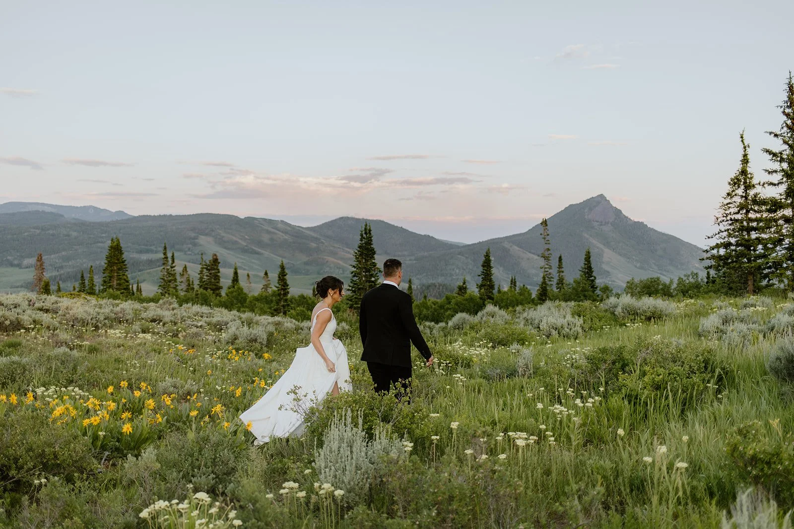 Bride in a white wedding gown and groom in a black tuxedo walking in a lush green meadow with wildflowers, with mountains and pine trees in the background, during sunset.