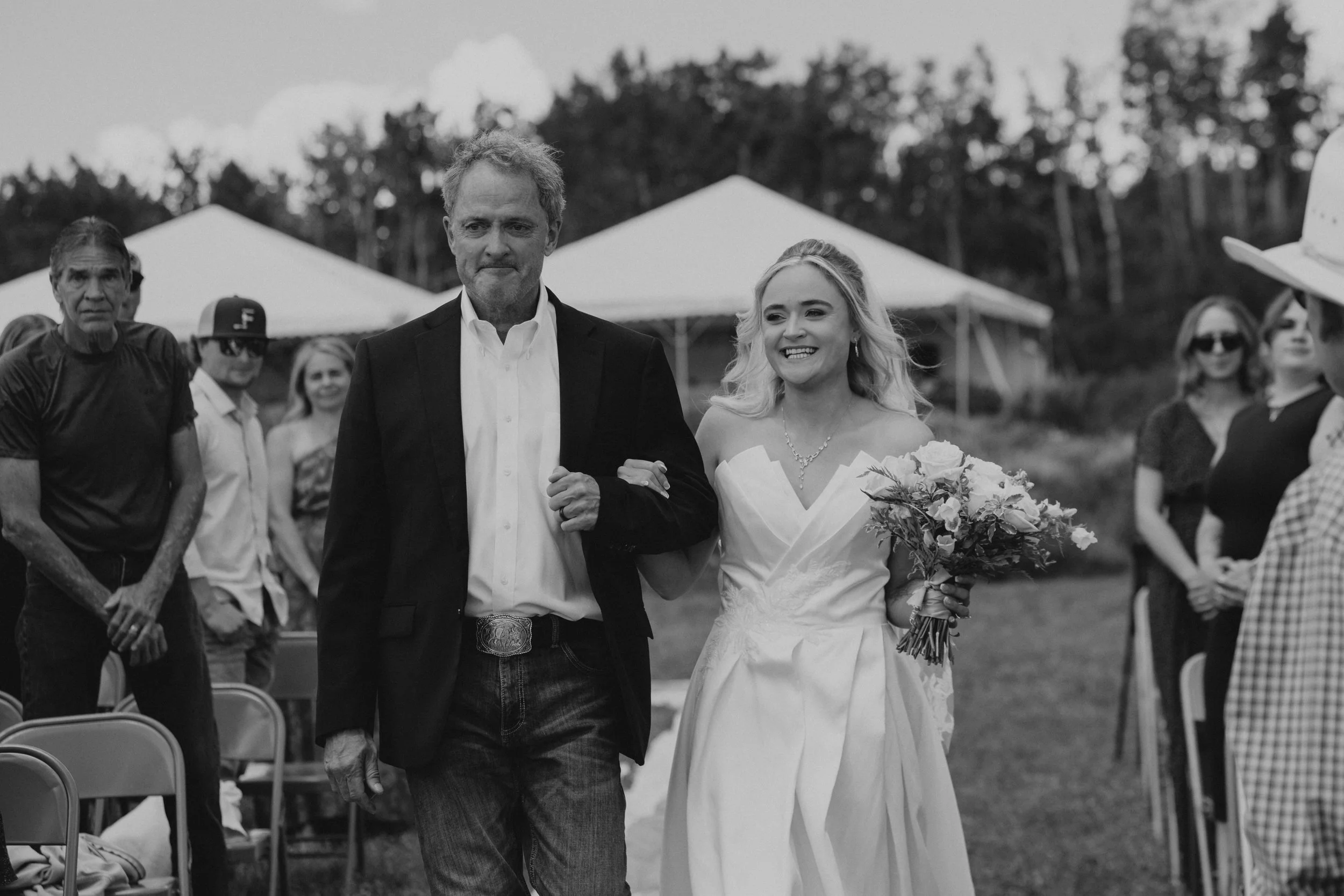 A black and white photo of a bride and an older man walking arm in arm at an outdoor wedding ceremony, with guests in the background and tents nearby.