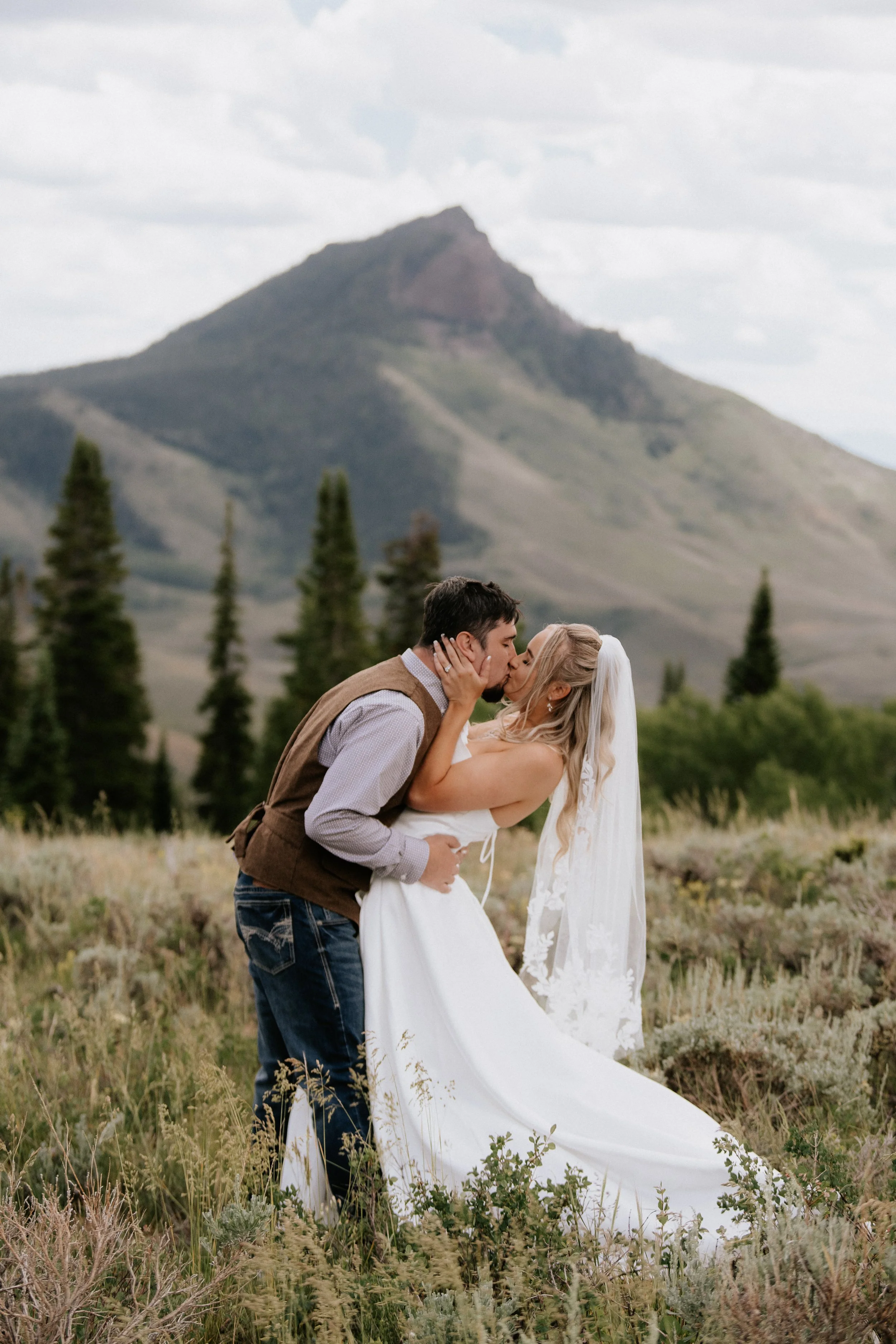 A newlywed couple sharing a kiss outdoors in a field with mountain scenery in the background. The groom holds the bride, who is wearing a white wedding dress, as they kiss.
