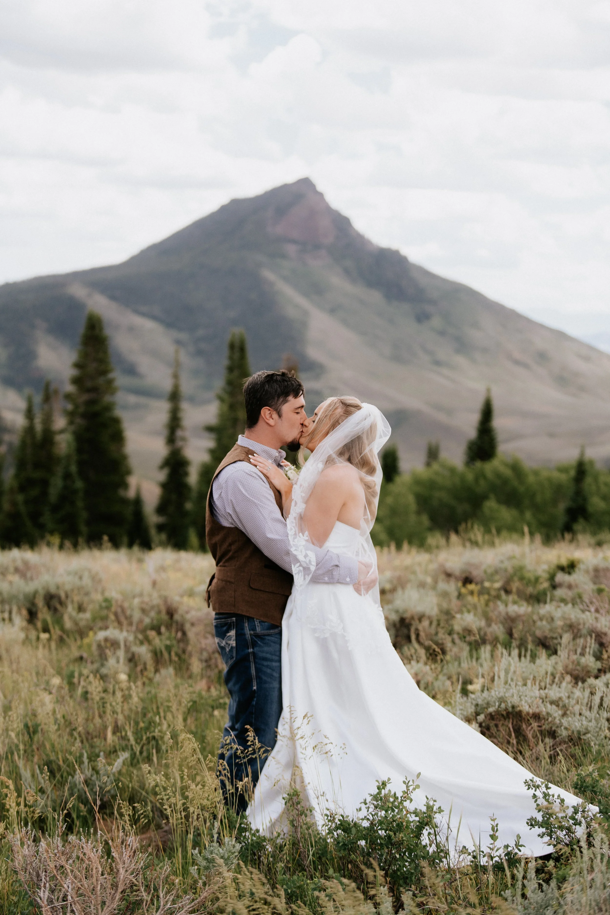A couple in wedding attire sharing a kiss in a grassy field with mountains and trees in the background.