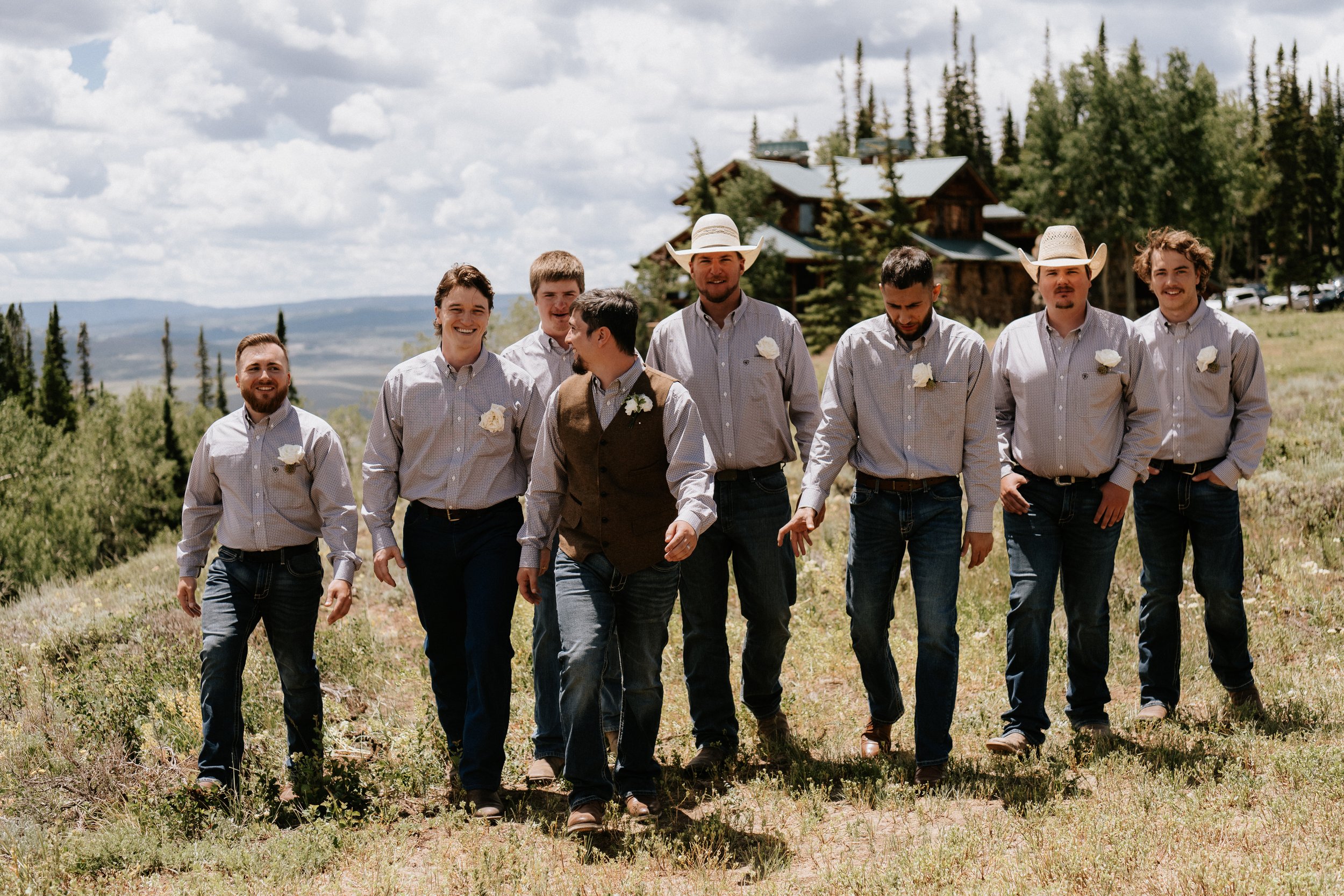A group of eight men walking outdoors in a rural, mountainous area with a wooden house in the background, dressed casually with some wearing cowboy hats.