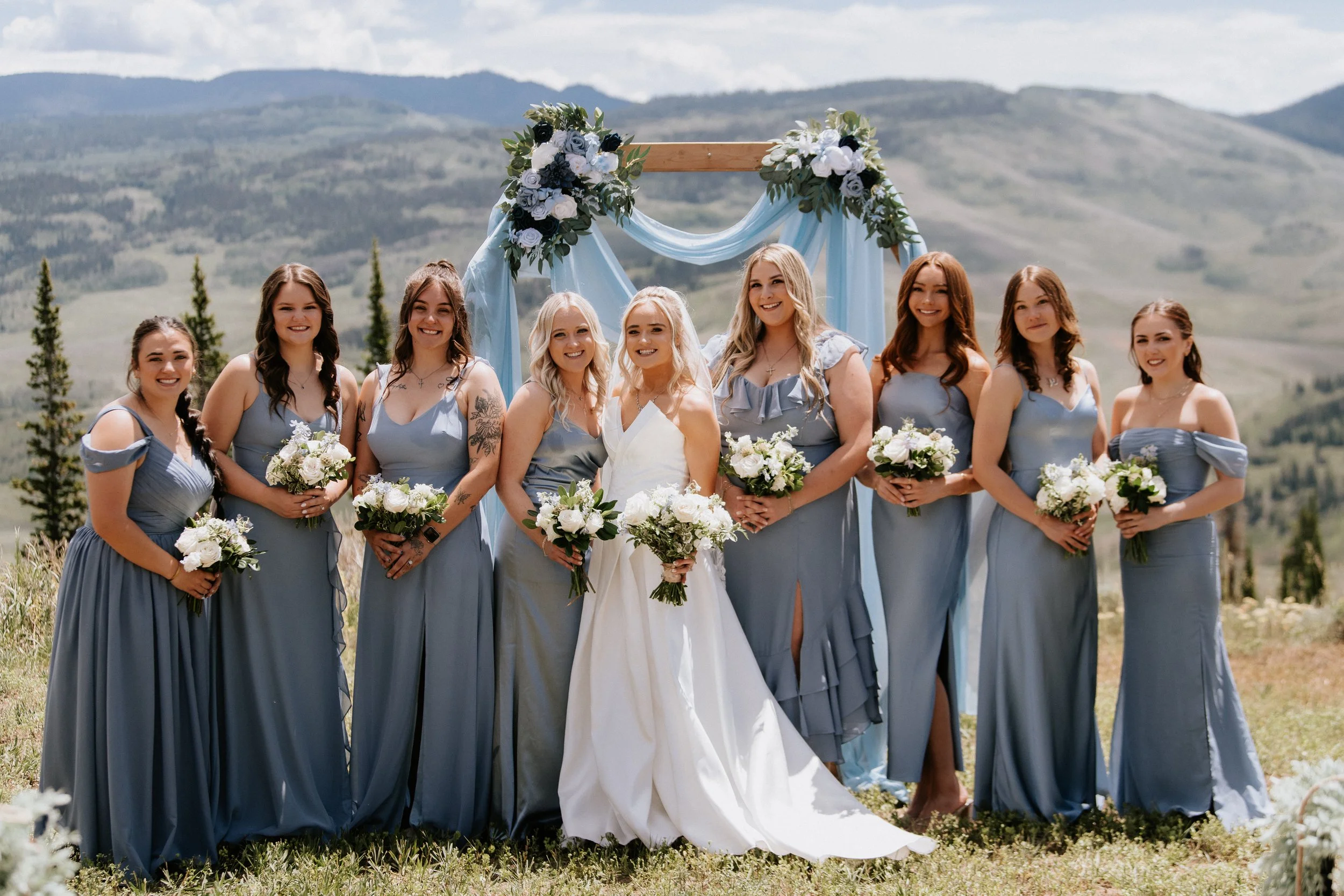 A group of women at a wedding outdoors in a mountainous area under a cloudy sky. The women are wearing pastel blue dresses, and the bride is in a white gown with blonde hair. They are holding white bouquets and standing in front of a decorated wooden