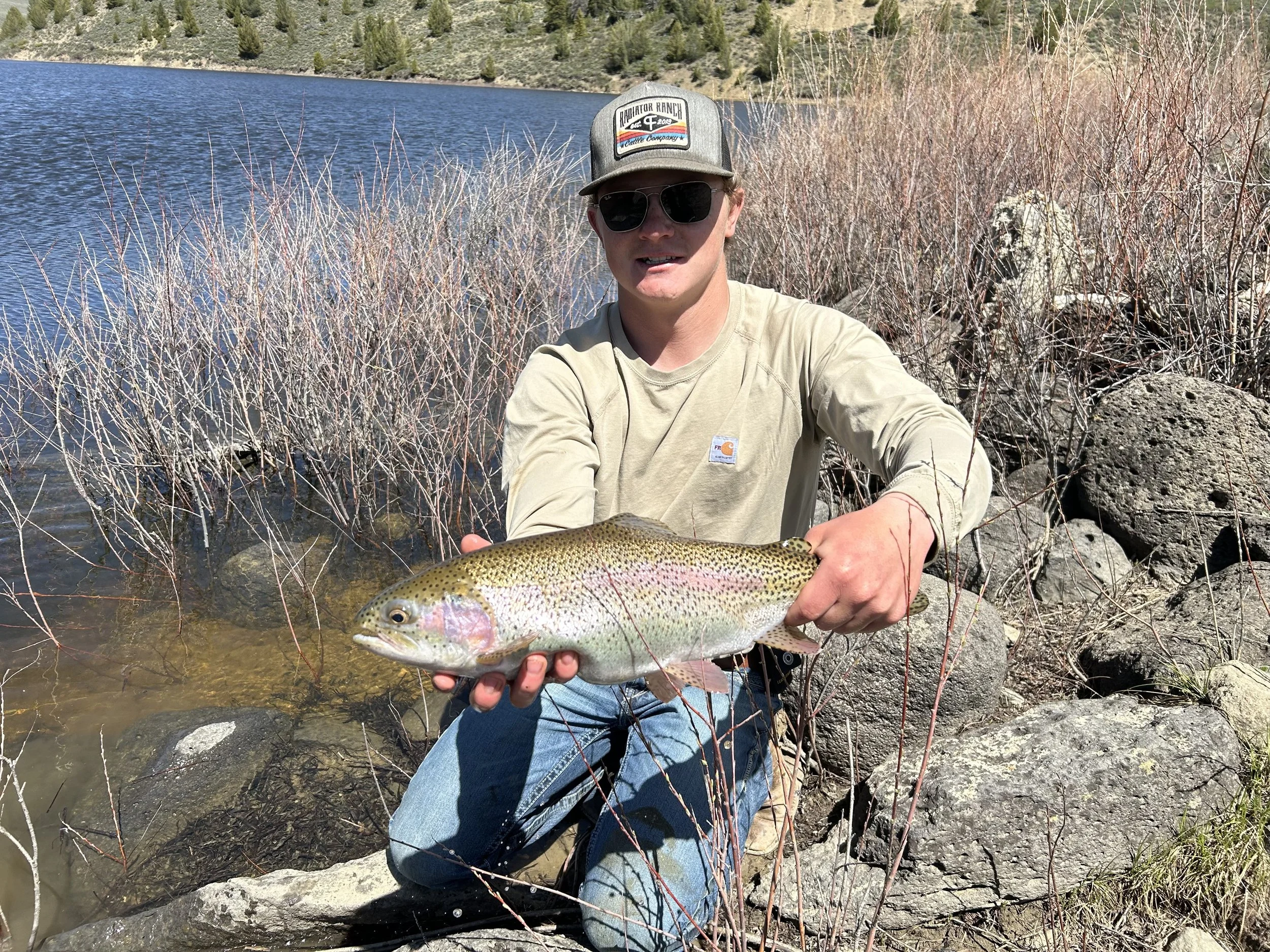Person wearing sunglasses and a cap holding a large rainbow trout by the lake shore, with rocks and dry bushes in the background.