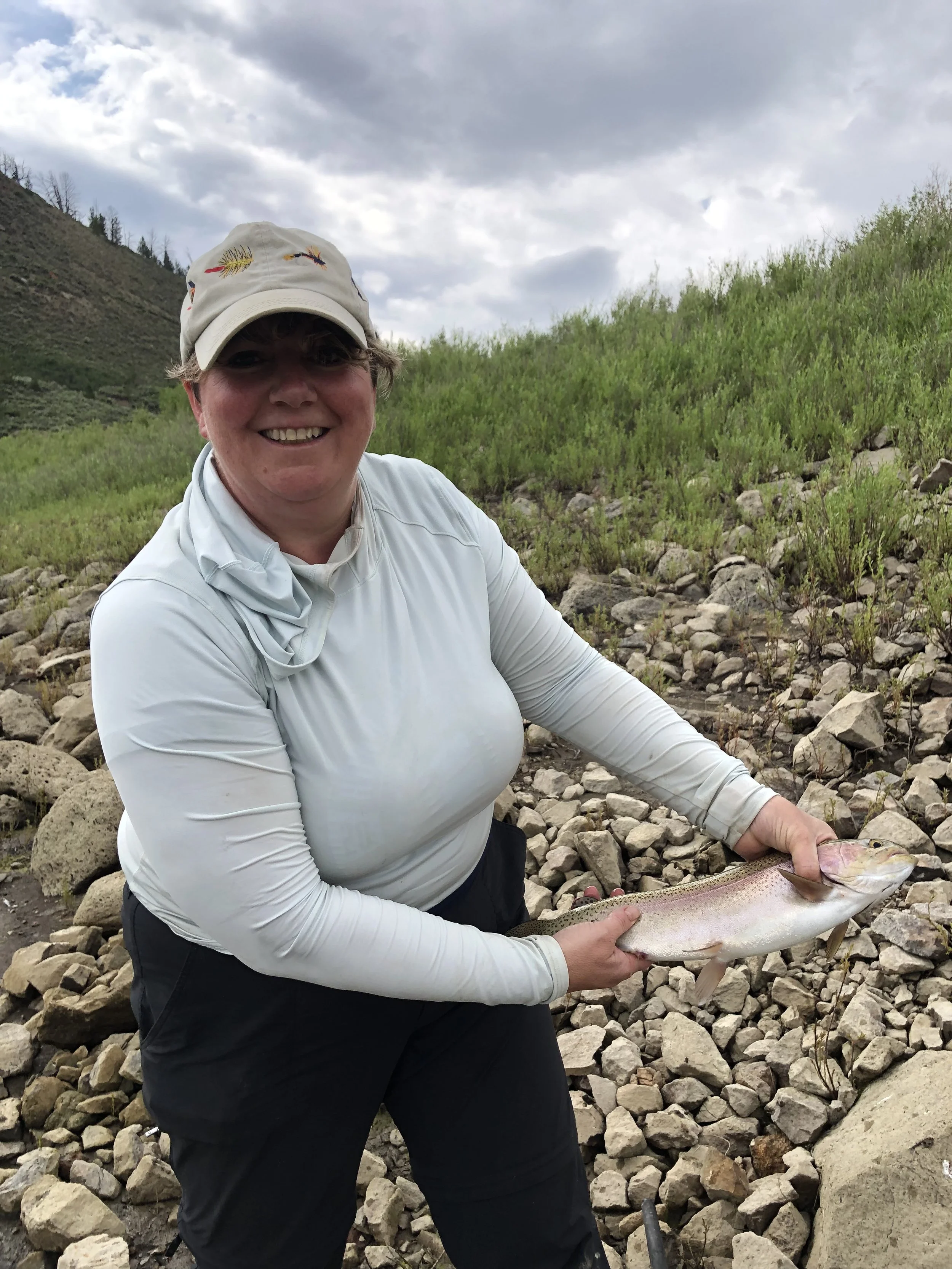 A woman smiling and holding a large fish outdoors on rocky terrain with green bushes and hills in the background, underneath a cloudy sky.