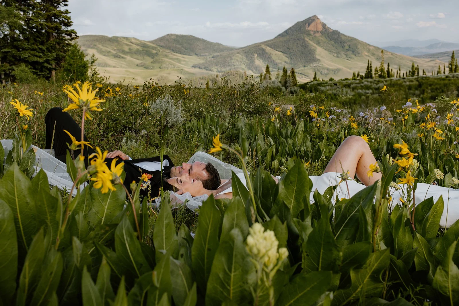 A couple lying in a field of yellow and purple wildflowers with mountains in the background, smiling and enjoying the outdoors.