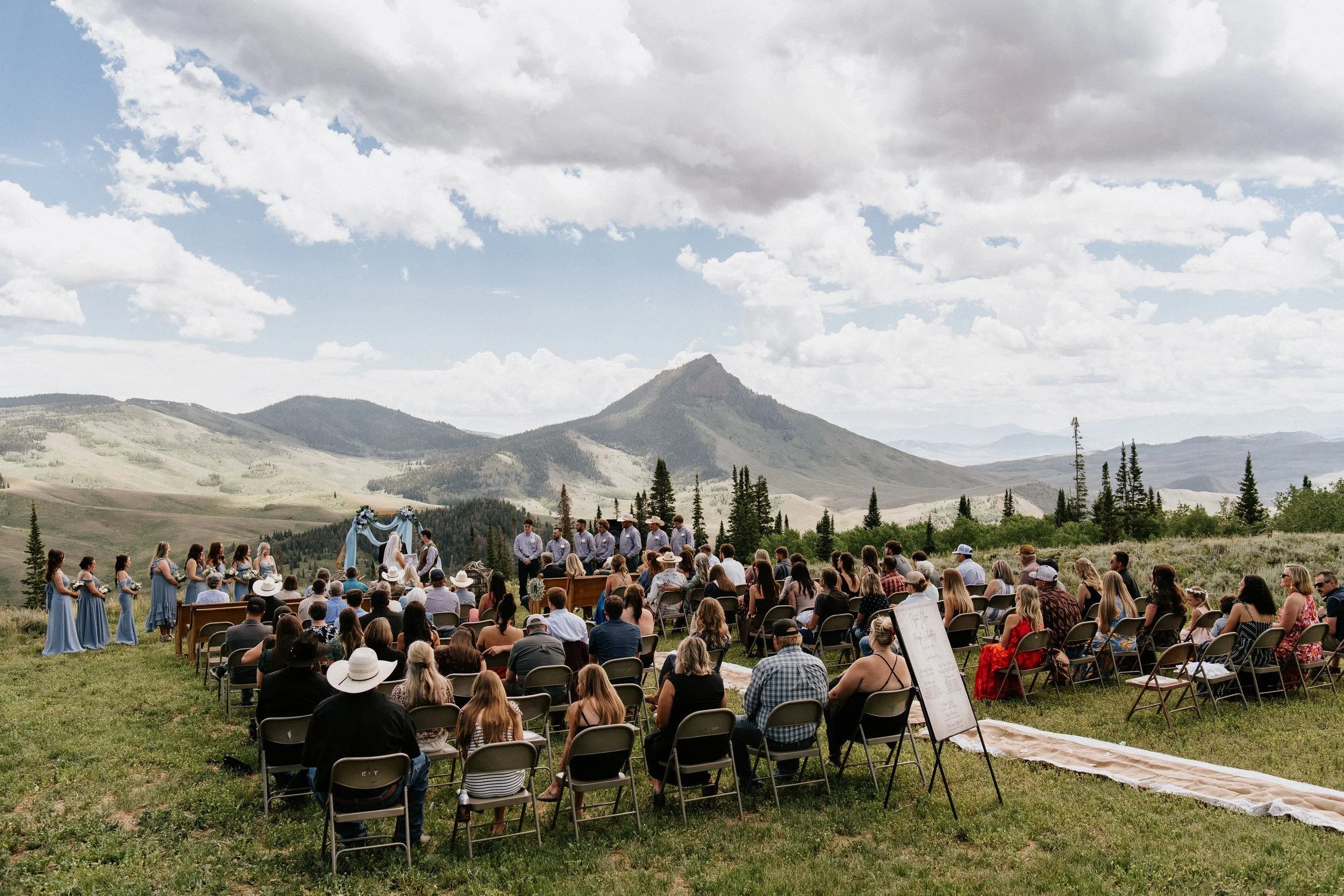 Outdoor wedding ceremony with guests seated on chairs, officiant and wedding party at the altar, set against mountainous landscape under cloudy sky.