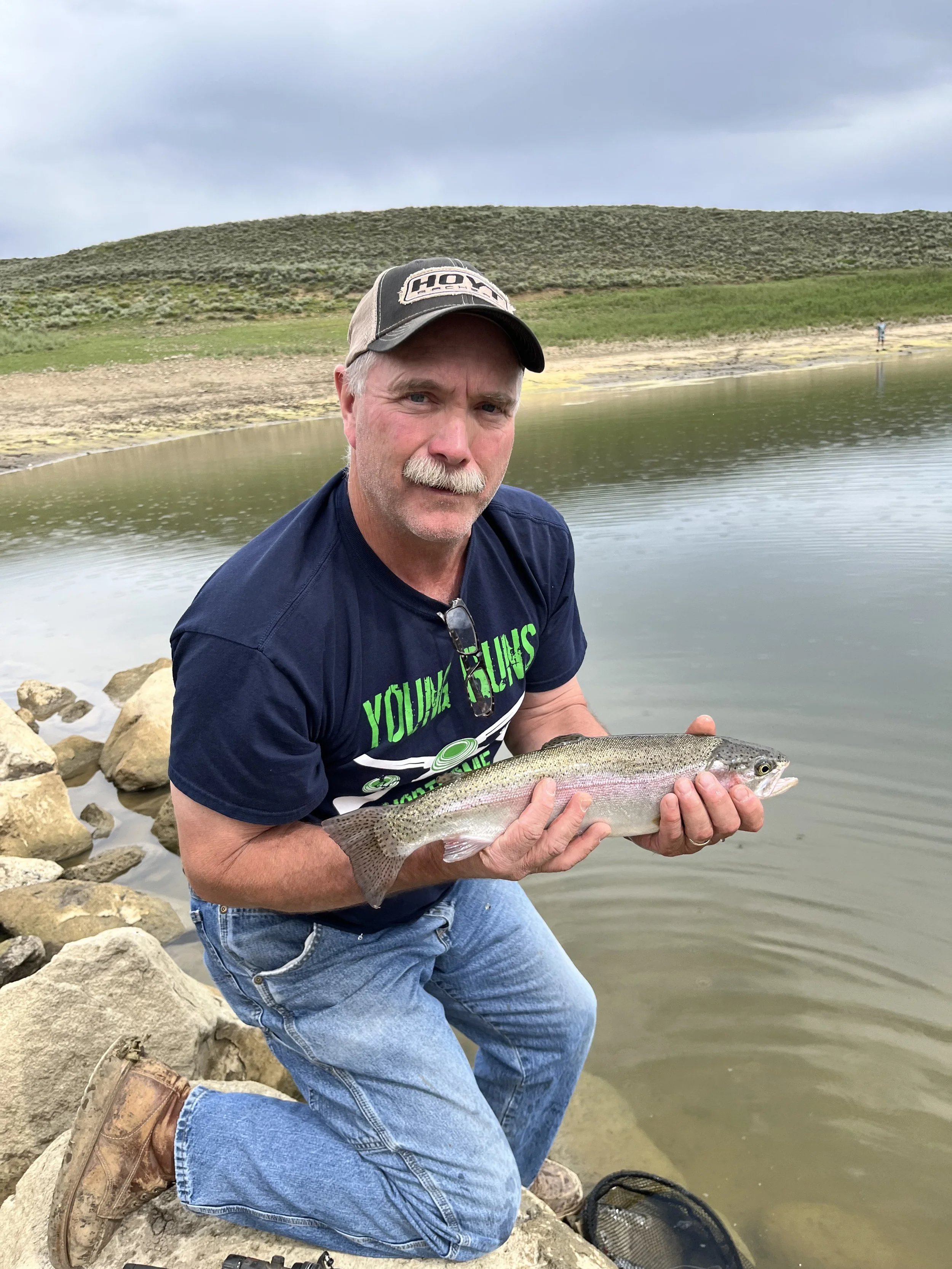 Man kneeling by a river holding a fish, with rocks and a grassy hill in the background under a cloudy sky.