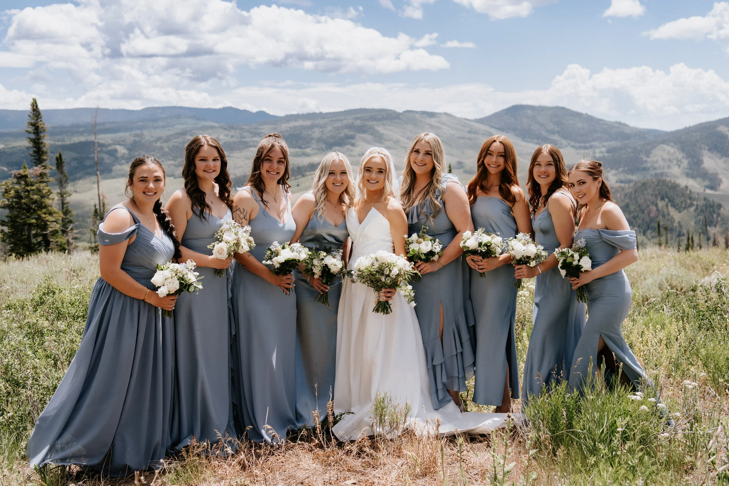 A group of nine women in blue bridesmaid dresses and one bride in a white wedding dress standing outdoors in a field with mountains and trees in the background, holding bouquets, on a sunny day with scattered clouds.