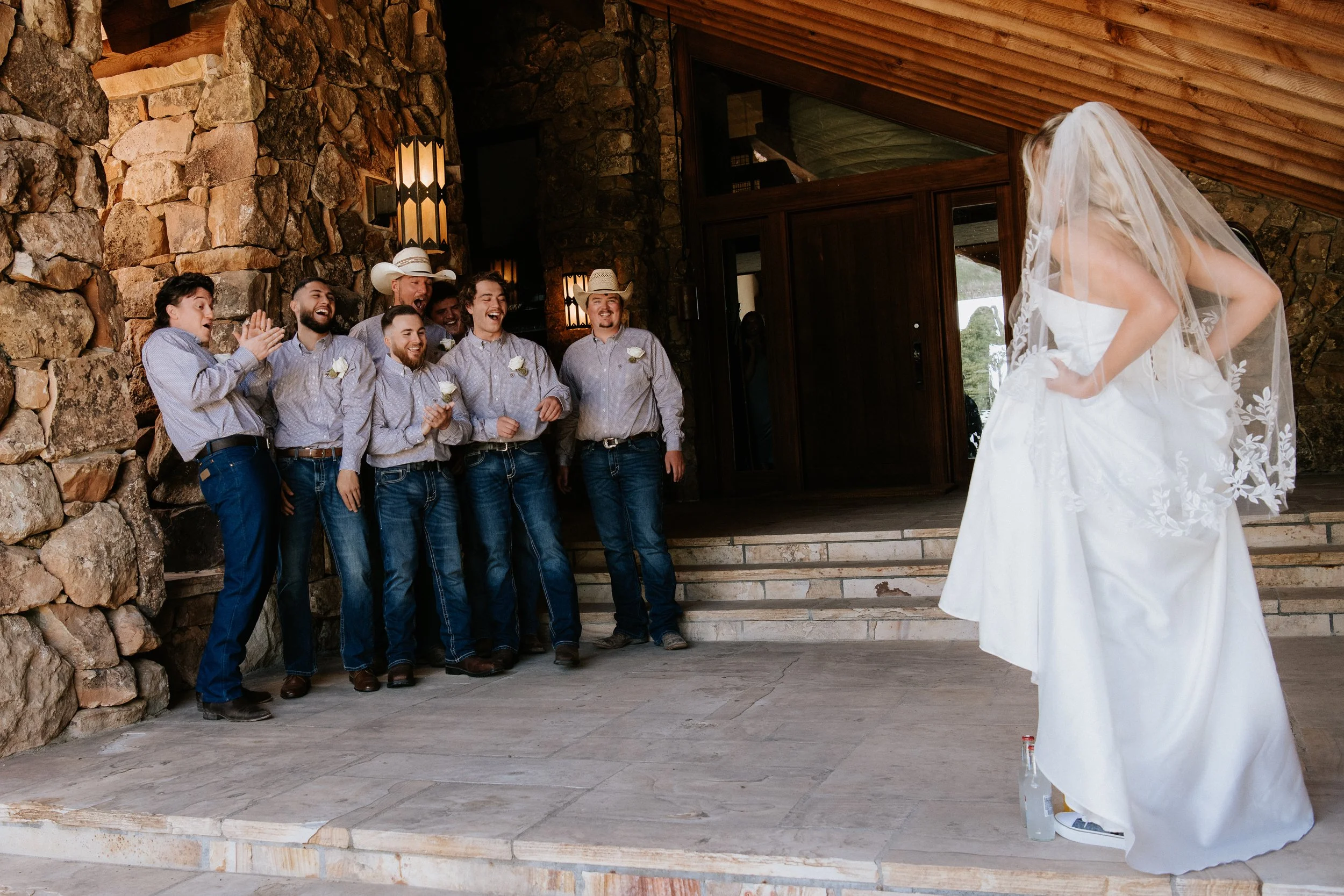 A bride in a white wedding dress looking at a group of men, who are laughing and clapping as they look at the bride. The men are wearing cowboy hats and dress shirts, standing on stone steps outside a rustic building with stone walls and wooden ceili