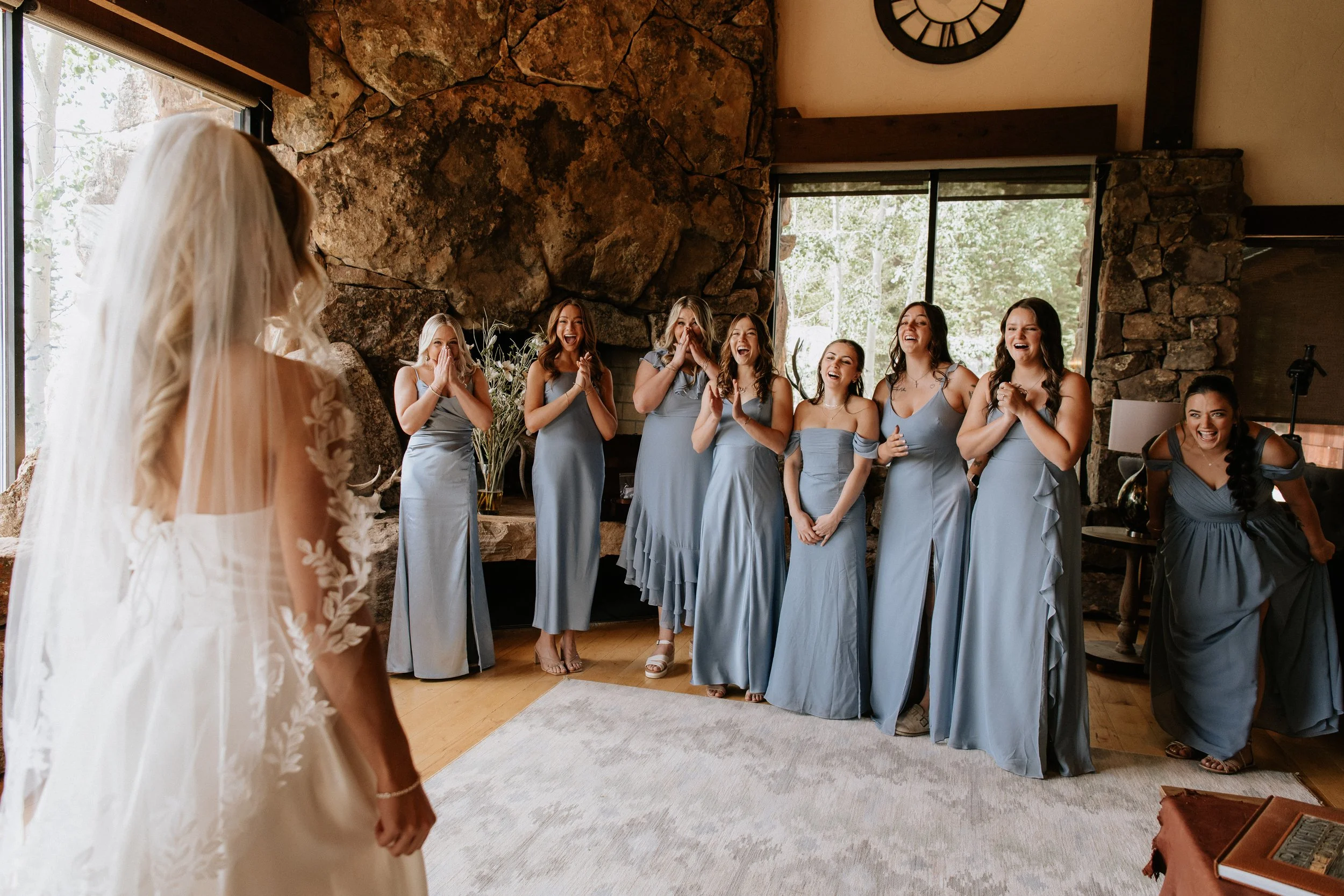 A bride in a wedding dress is standing in front of a group of bridesmaids in matching light blue dresses, all of whom are smiling and laughing inside a room with large windows, a stone wall, and a wooden ceiling.