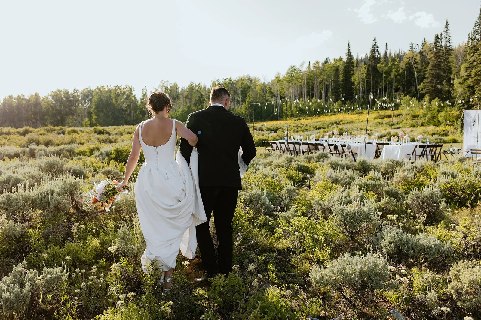 Bride and groom walking through a field towards an outdoor wedding reception setup with long tables and chairs, surrounded by greenery and trees.