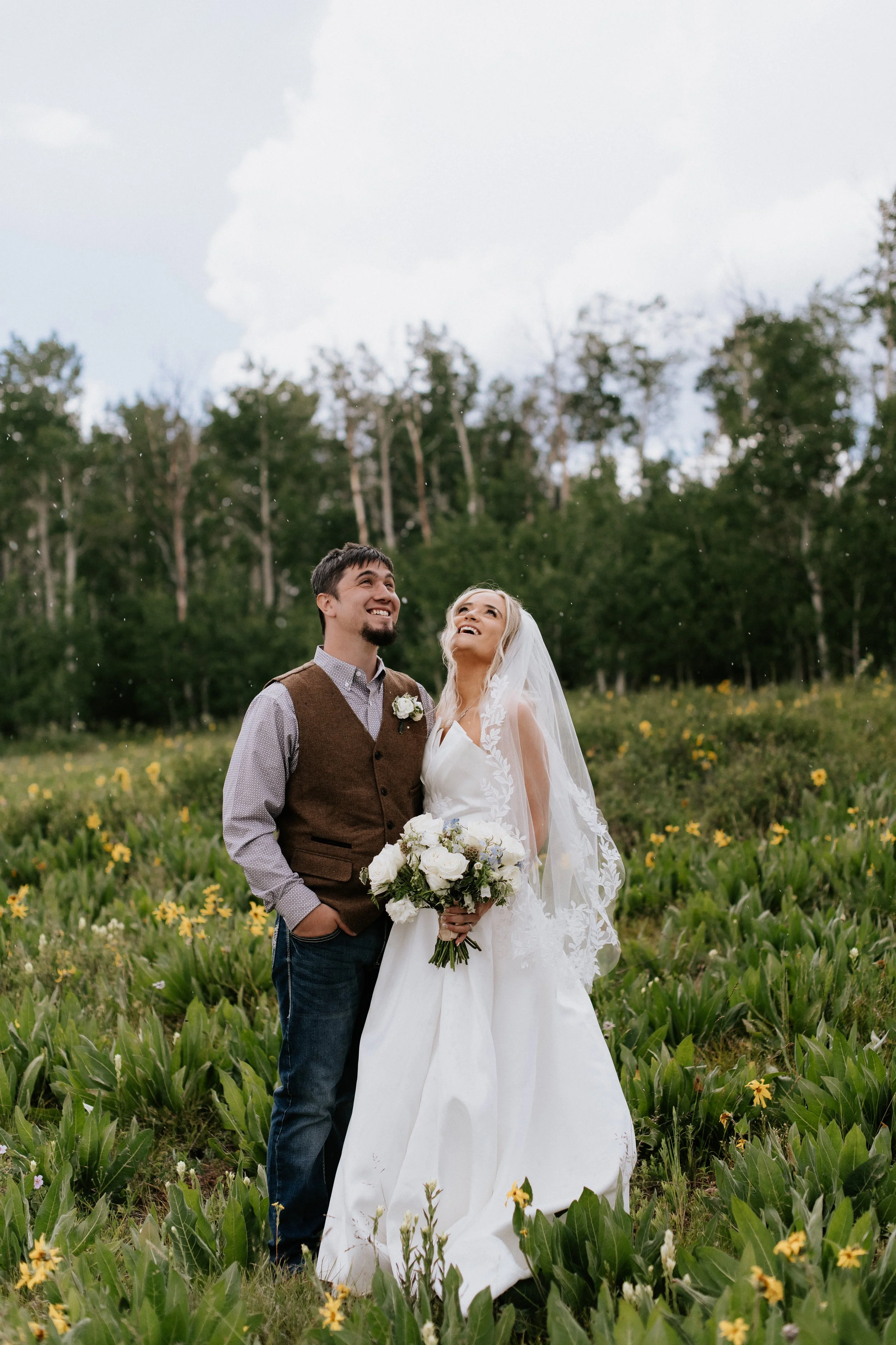 A bride and groom stand closely together in a lush field of green plants and yellow flowers outdoors, smiling and looking up, with a backdrop of trees and a cloudy sky.