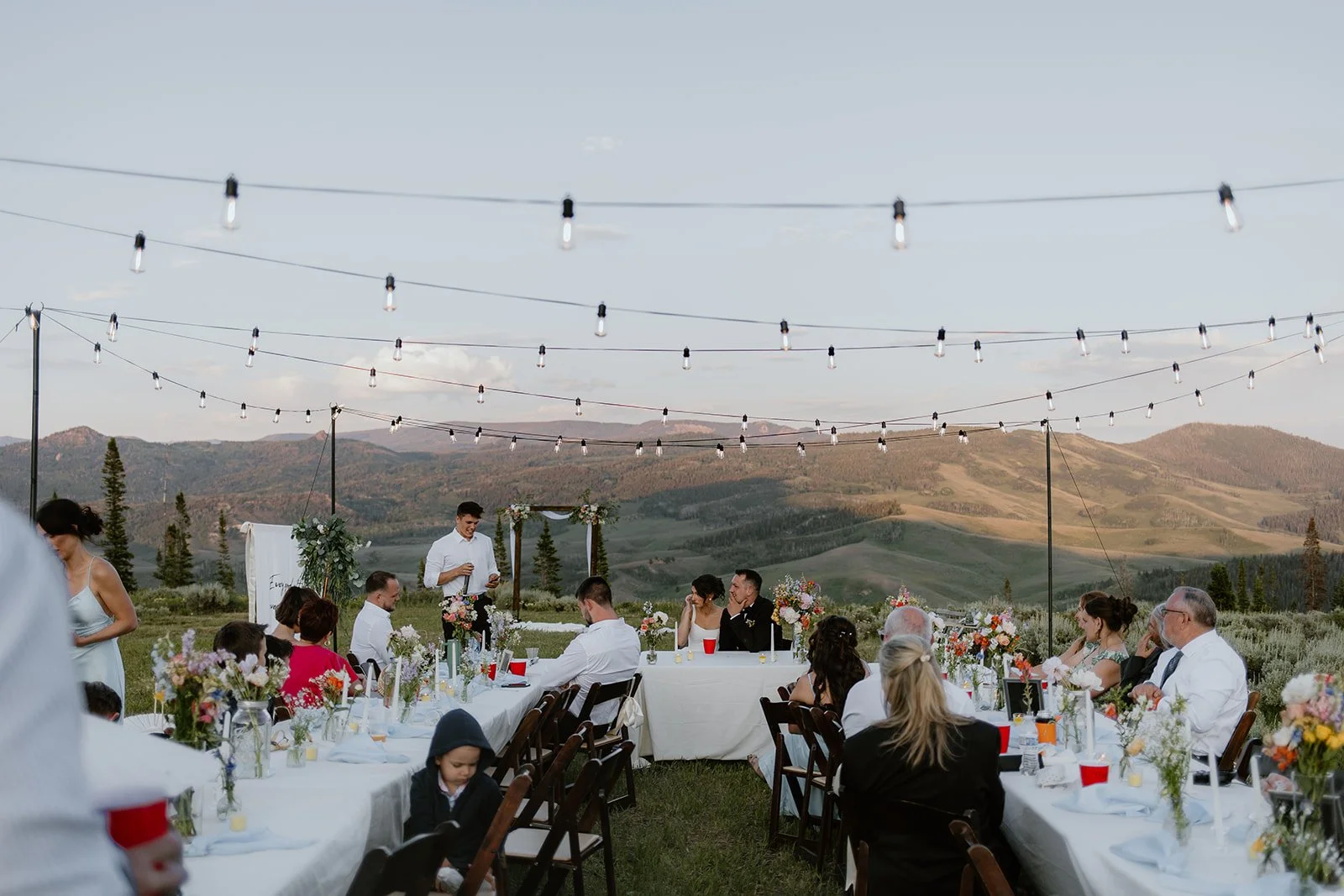 Outdoor wedding reception with guests seated at long tables decorated with flowers, in a scenic mountainous setting under string lights, during late afternoon or early evening.