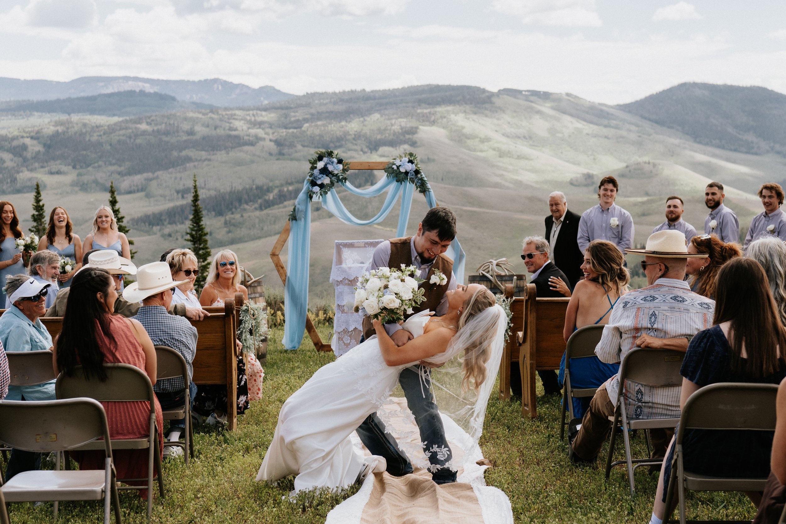 A bride and groom exchange vows at an outdoor wedding ceremony with mountains in the background, guests seated on chairs, and wedding decorations including floral arrangements and drapery.