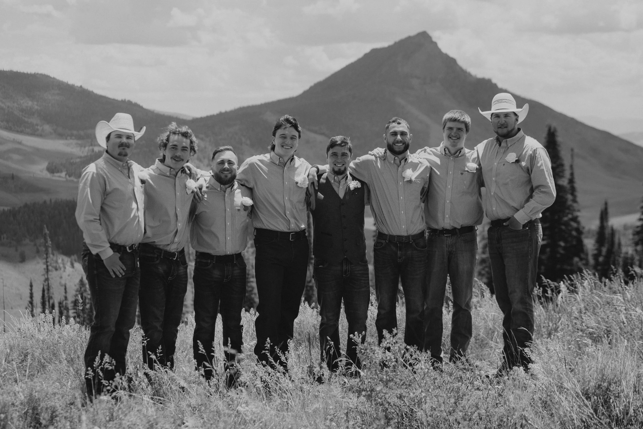 Group of nine men standing outdoors in a row, smiling, with mountains in the background. Most are wearing collared shirts, some with hats, and have boutonnières on their shirts, suggesting a wedding or celebration.