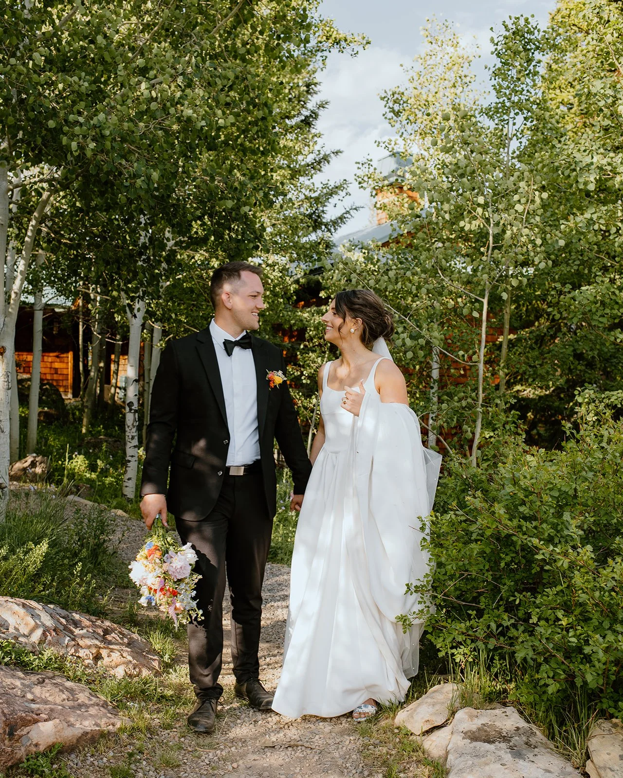 A bride and groom are standing outdoors, holding hands, surrounded by trees and greenery, smiling at each other.